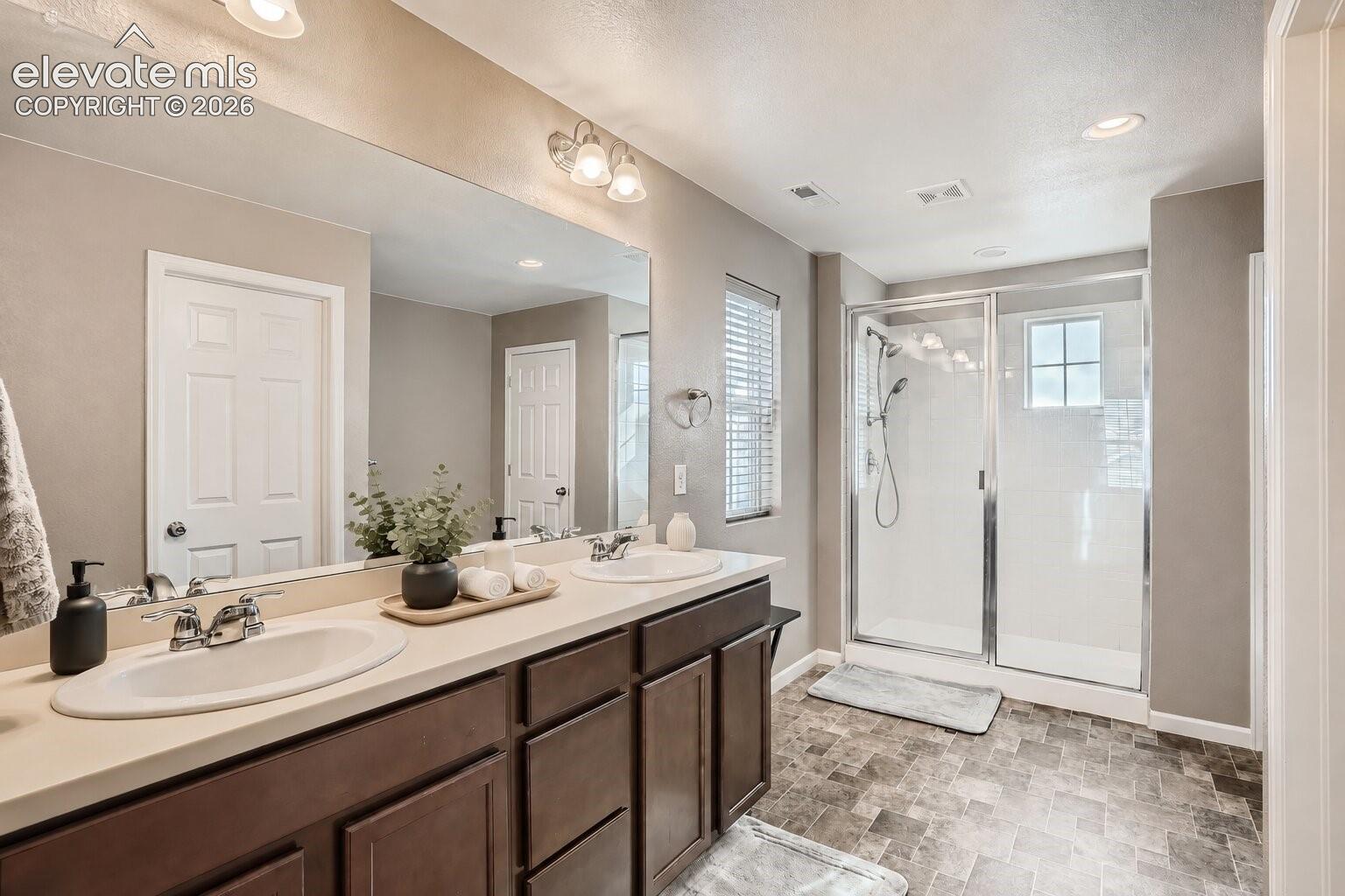 Dual-sink vanity with a solid-surface countertop, full-width wall mirror, dark-toned cabinetry, and walk-in shower with a glass enclosure