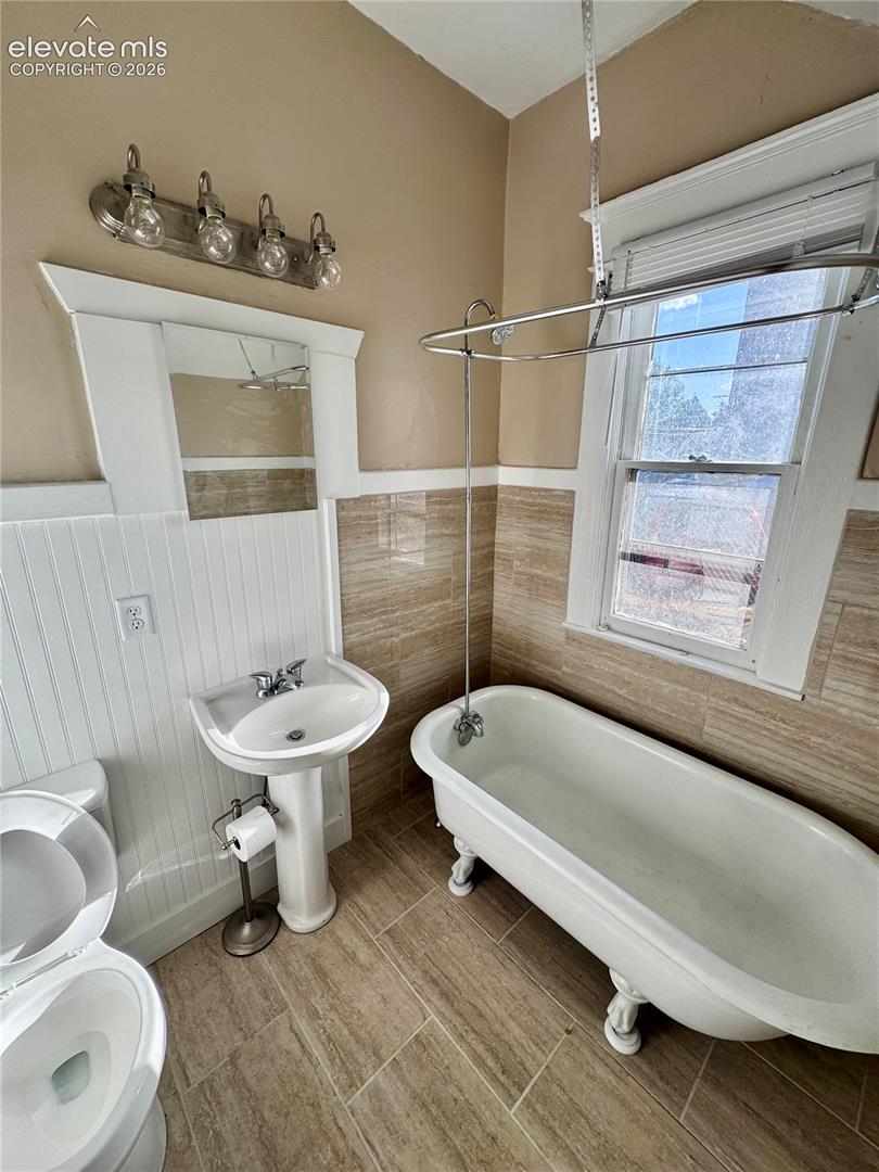 Pedestal sink, clawfoot tub, window with blinds, tile flooring, and wall-mounted vanity lighting