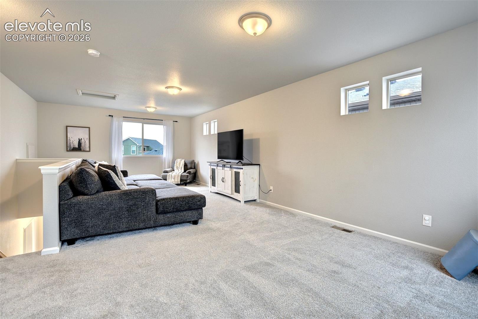 Open interior space featuring light-colored wall-to-wall carpeting, neutral wall tones, multi-level ceiling lighting, and multiple windows including high-placement square casements