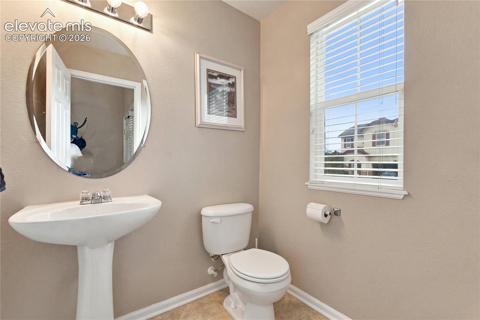 Oval wall-mounted mirror, pedestal sink with chrome faucet, multi-light fixture, dual-pane window with integrated blinds, and neutral wall tones