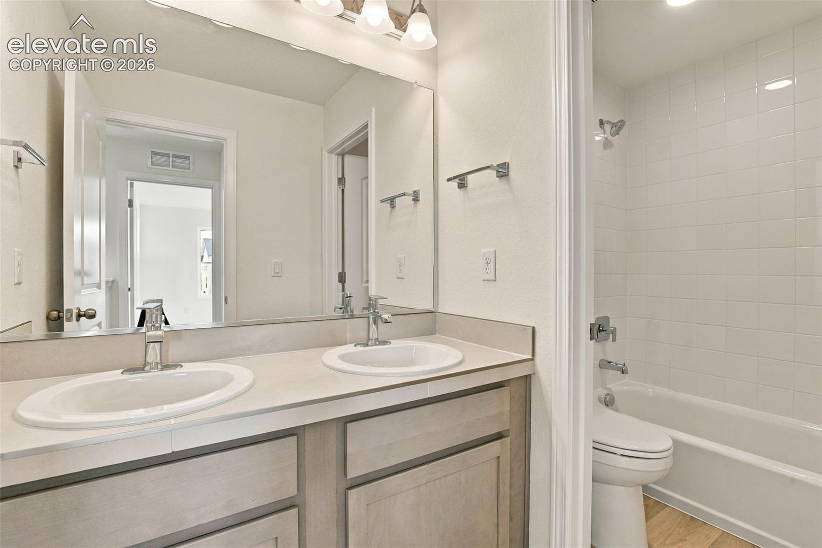 Dual-sink vanity with neutral countertop, full-width mirror, light-finish cabinetry, and tiled shower-tub combination