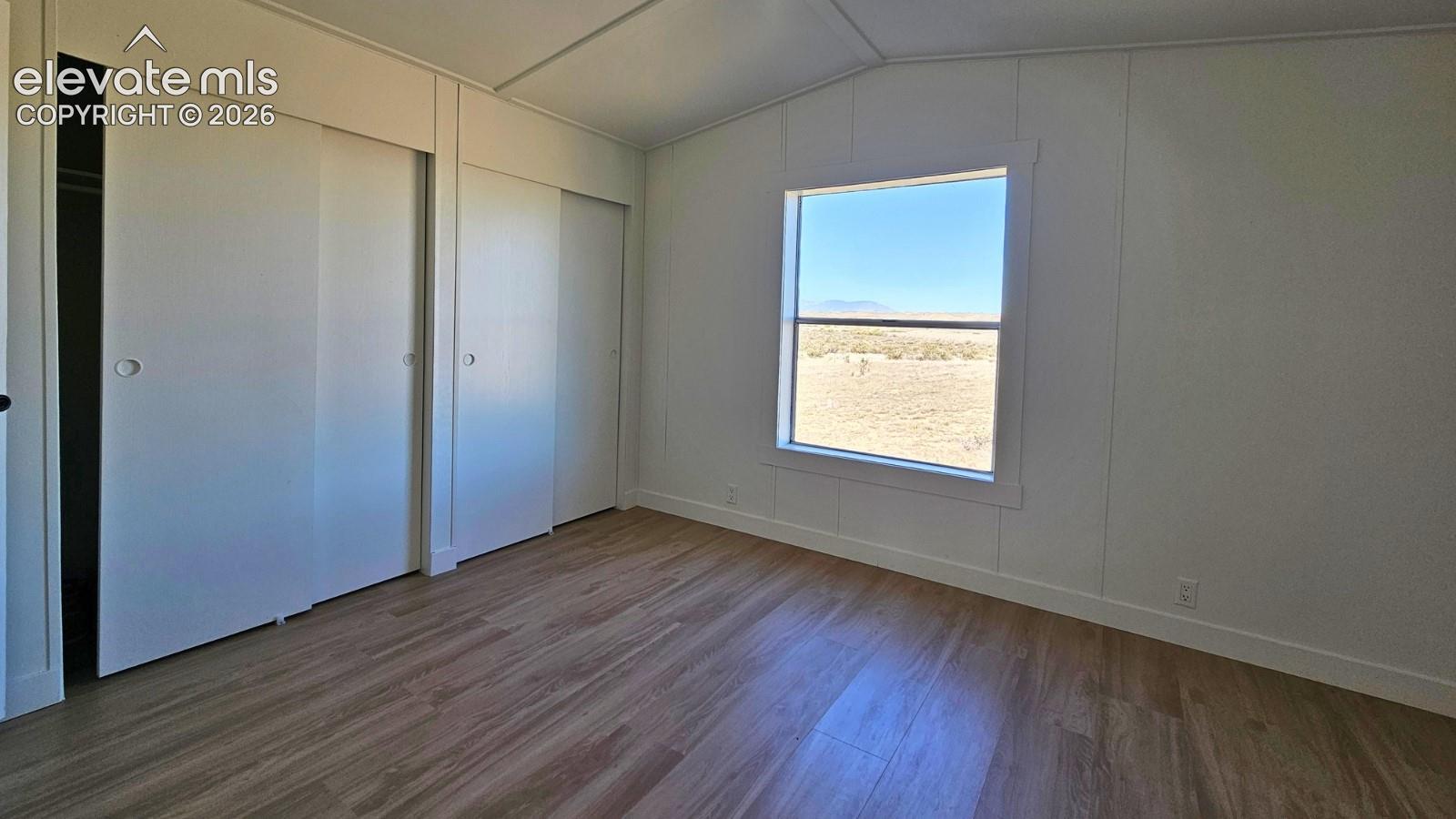 Interior space featuring wood-finish flooring, vaulted ceilings, neutral wall paneling, and dual sliding-door closets, with a window framing the exterior landscape