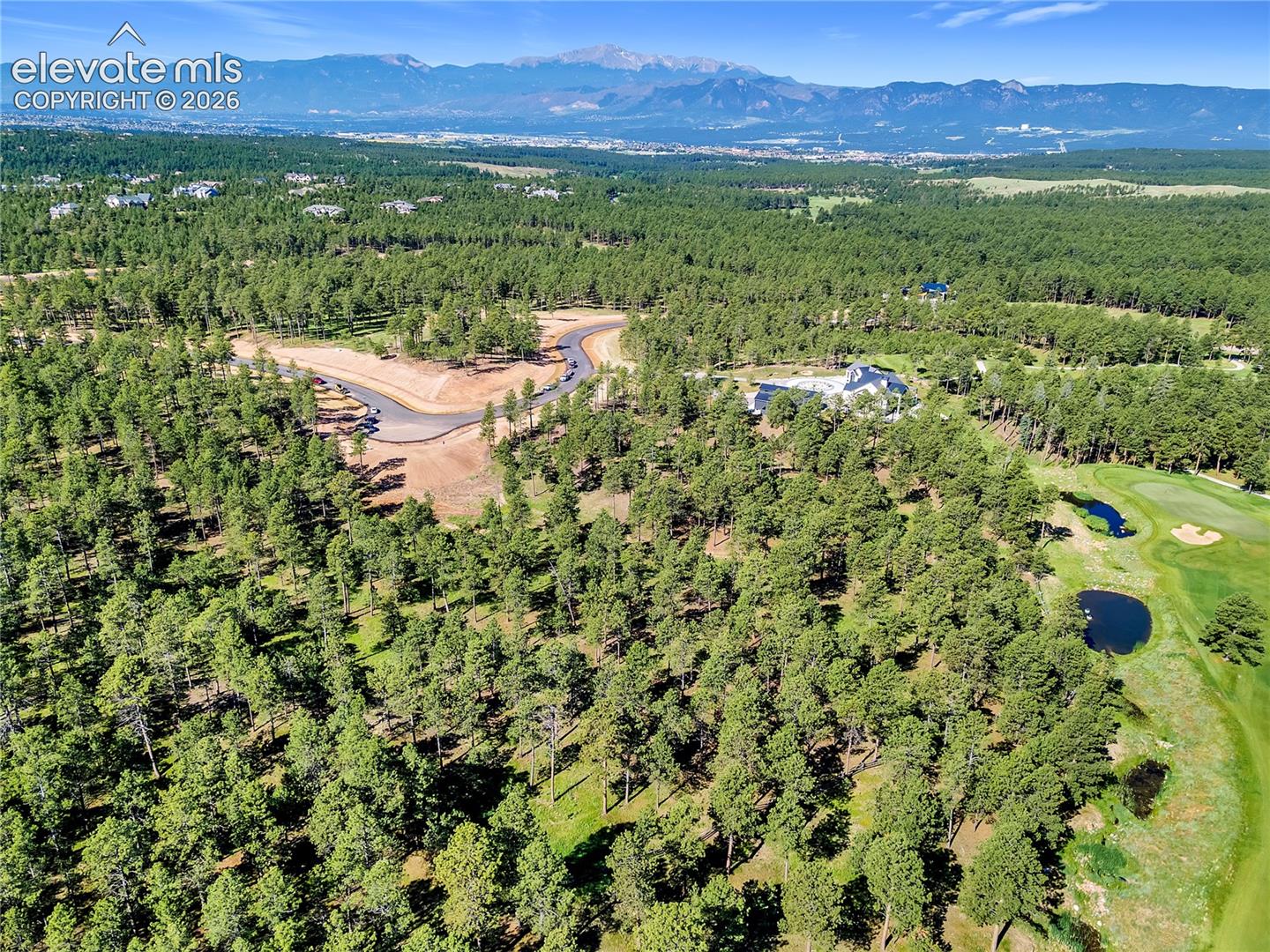 Aerial perspective of a large residential lot featuring dense pine forest, proximity to a golf course with water features, and mountain range scenery