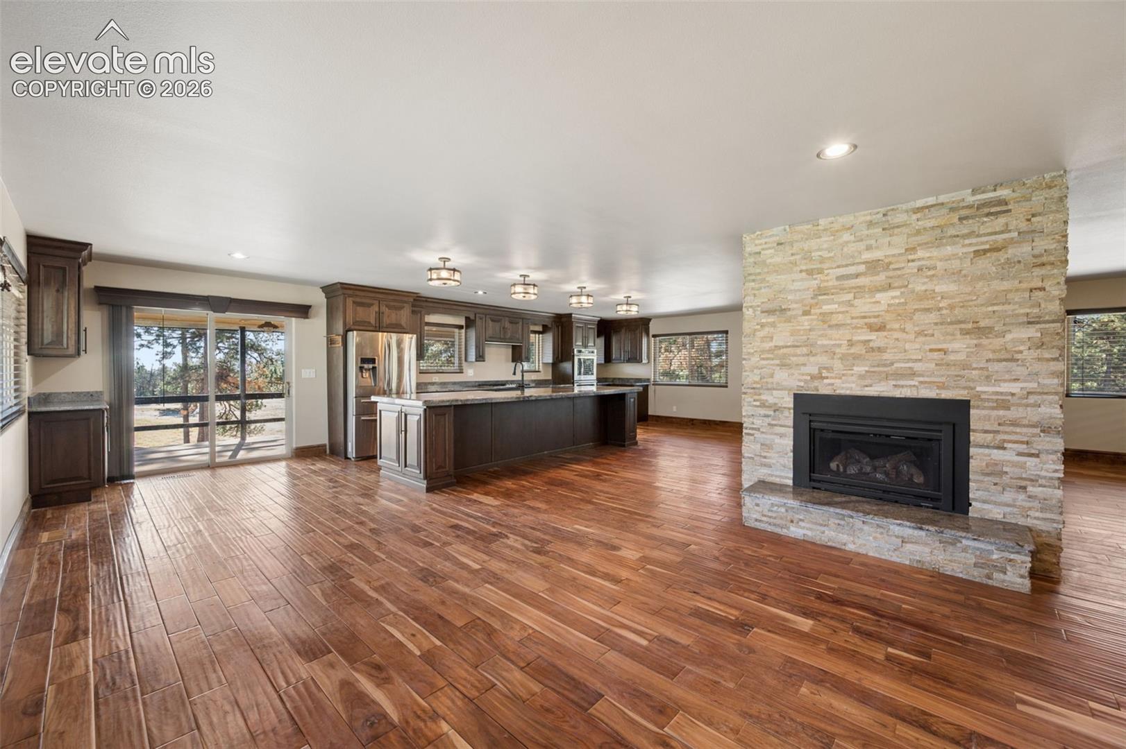 Wood-finish flooring, floor-to-ceiling stone-veneer fireplace with a built-in firebox, dark cabinetry, stone-surface kitchen island, and sliding glass door with exterior access