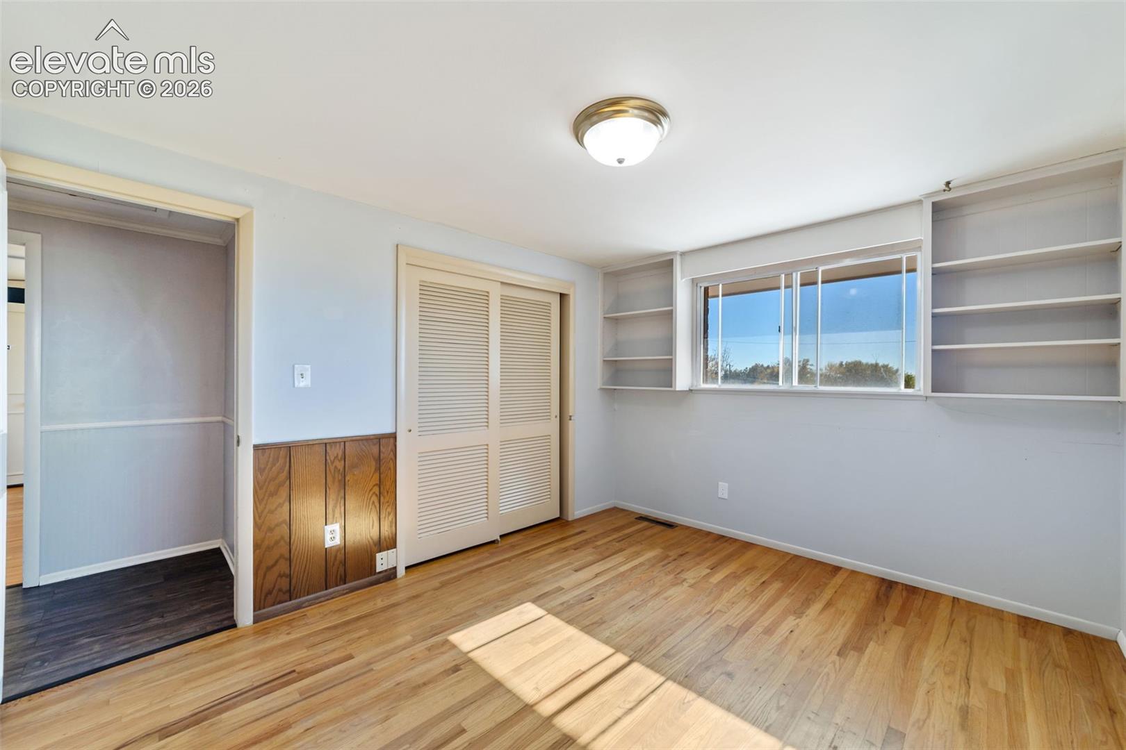 Room featuring wood-finish flooring, a window with exterior visibility, built-in shelving, a flush mount ceiling light, and louvered closet doors