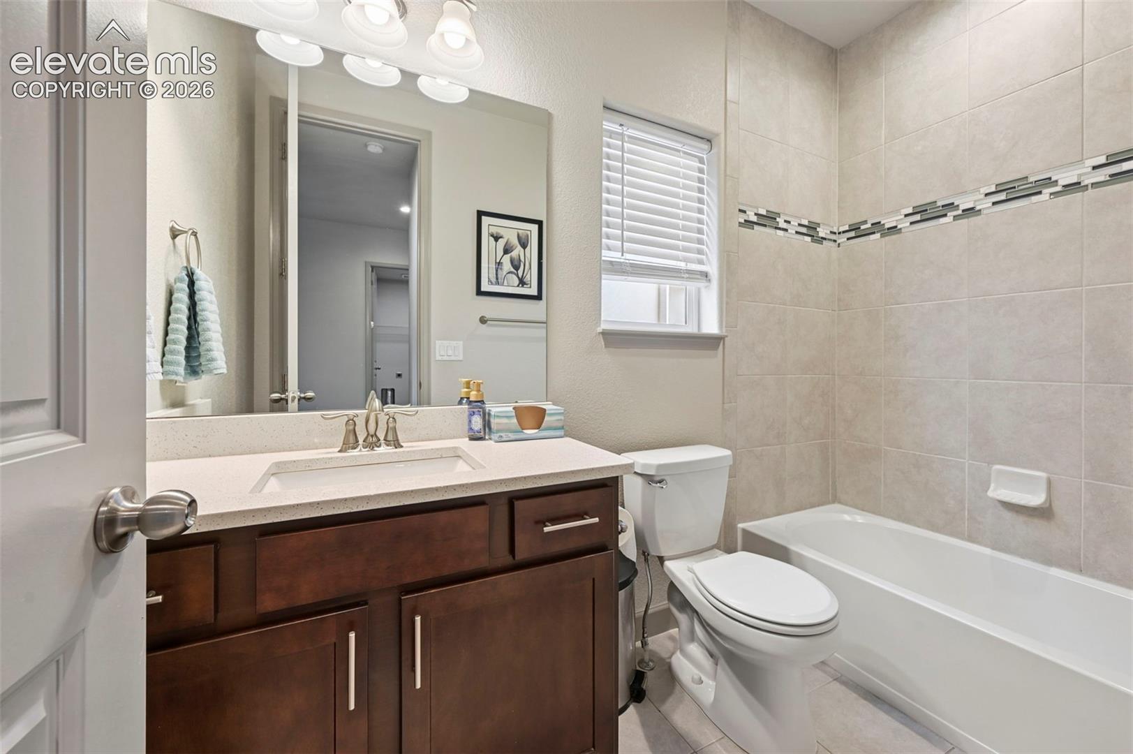 Dark wood-finish cabinetry, solid-surface countertop with an integrated sink, full-width mirror, and tiled shower-tub surround with a decorative mosaic accent border