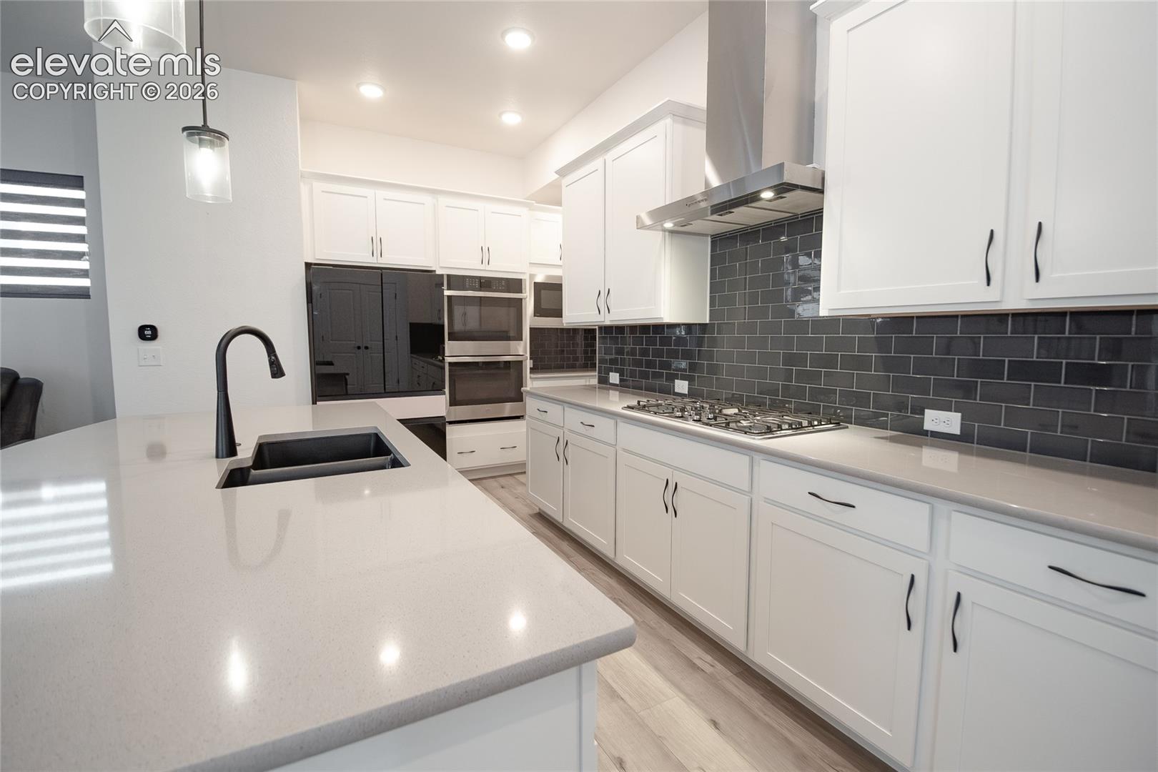 Kitchen featuring white shaker-style cabinetry, quartz countertops, a charcoal-toned subway tile backsplash, stainless steel double wall ovens, and a central island with an undermount sink