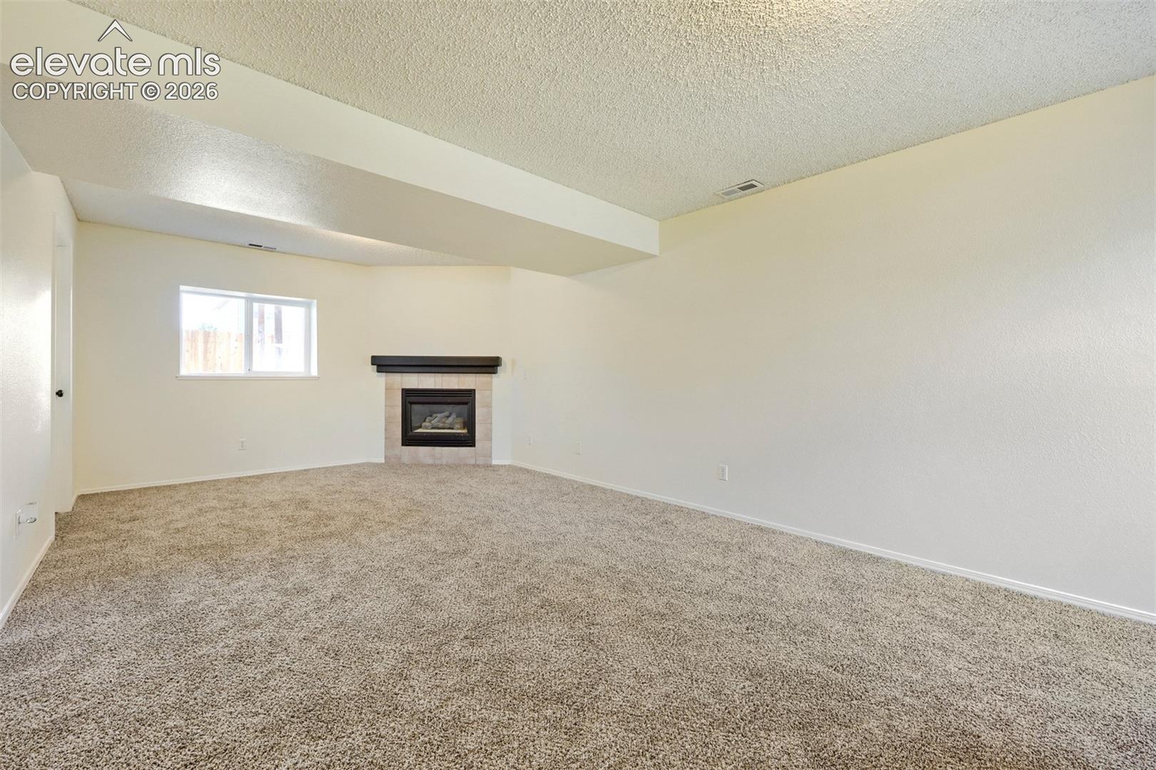 Corner fireplace with a dark wood mantel and tile surround, accompanied by a window with white trim, and light-toned carpet flooring
