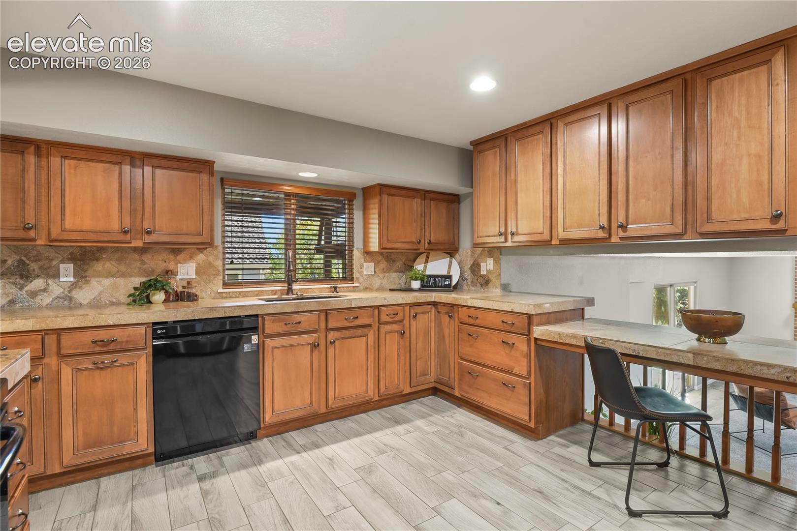 Kitchen featuring custom wood cabinetry, stone countertops, stone tile backsplash, wood-finish tile flooring, and a built-in countertop desk area