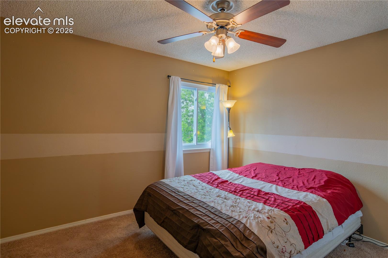 Textured ceiling, ceiling fan with integrated lighting, window with white curtain, tan painted walls, and wall-to-wall carpeting