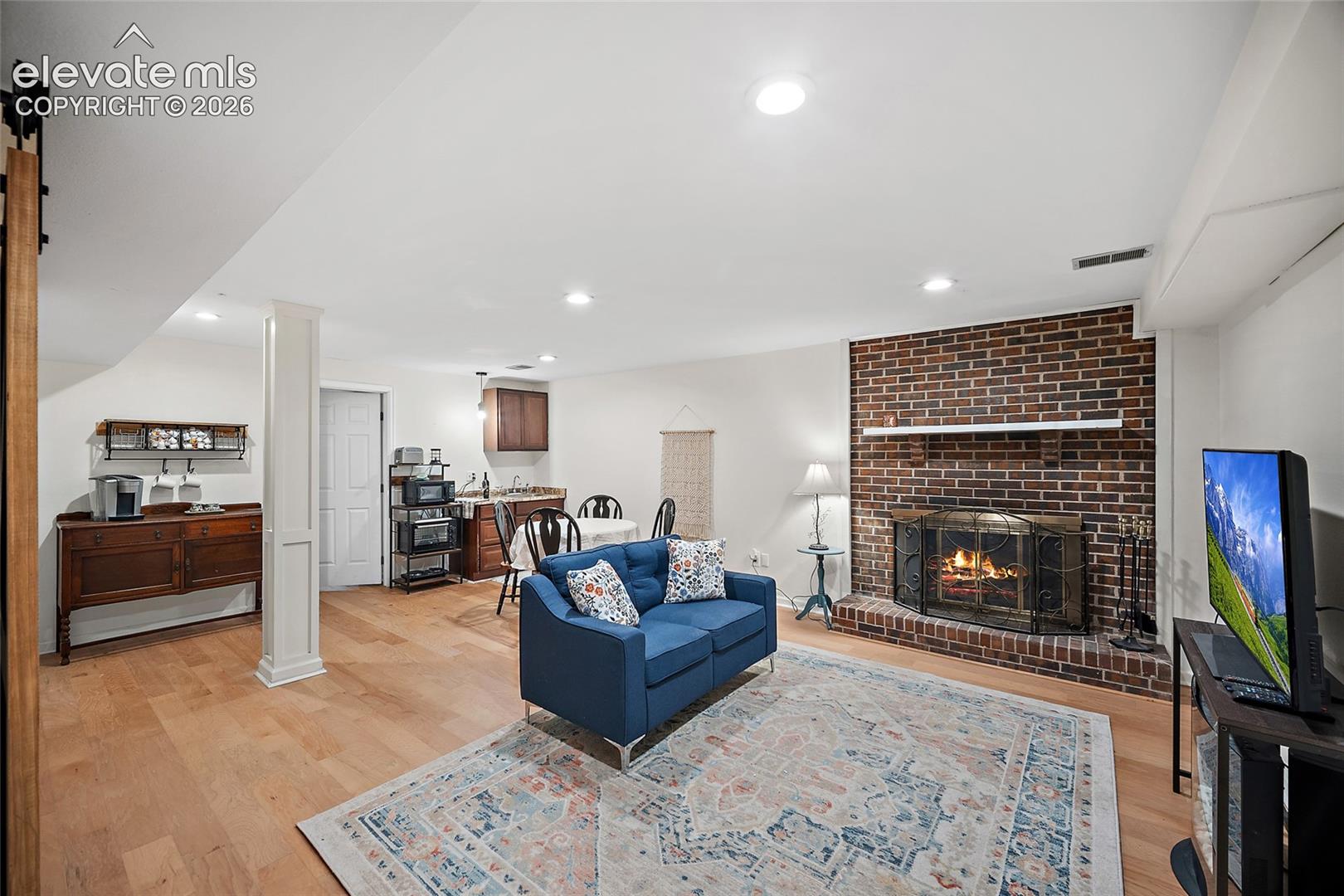 Open-concept living space featuring wood-finish flooring, a floor-to-ceiling brick fireplace with a mantel, recessed lighting, and a built-in kitchen kitchenette