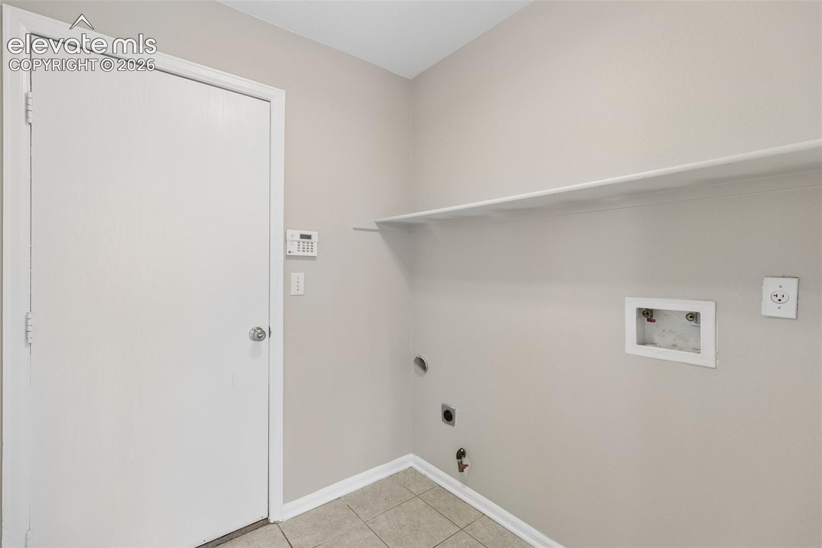 Utility space featuring neutral wall paint, tiled flooring, an overhead built-in storage shelf, and dedicated water and electrical hookups