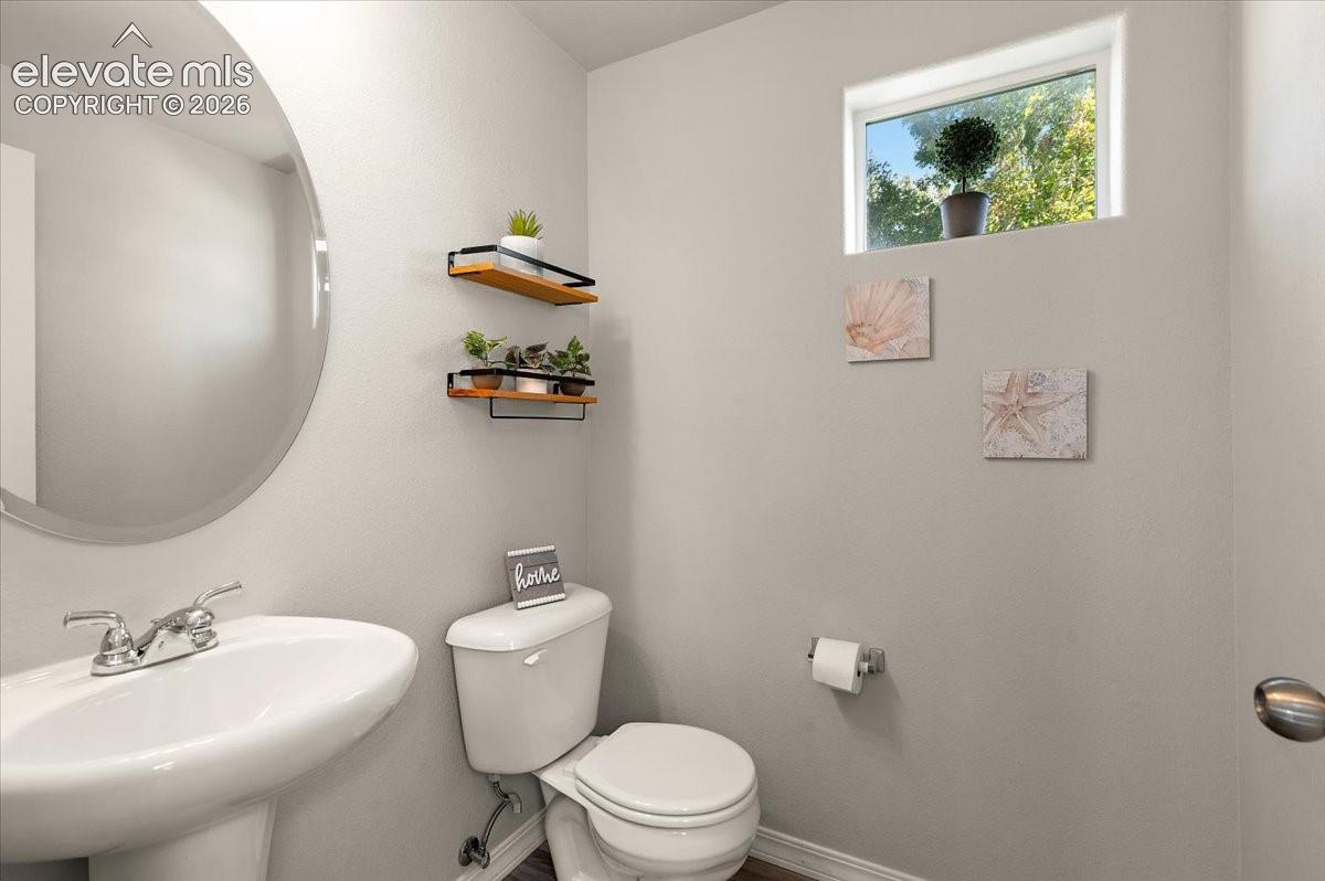 Pedestal sink with chrome faucet, circular wall mirror, wall-mounted shelving, privacy window, and wood-finish flooring