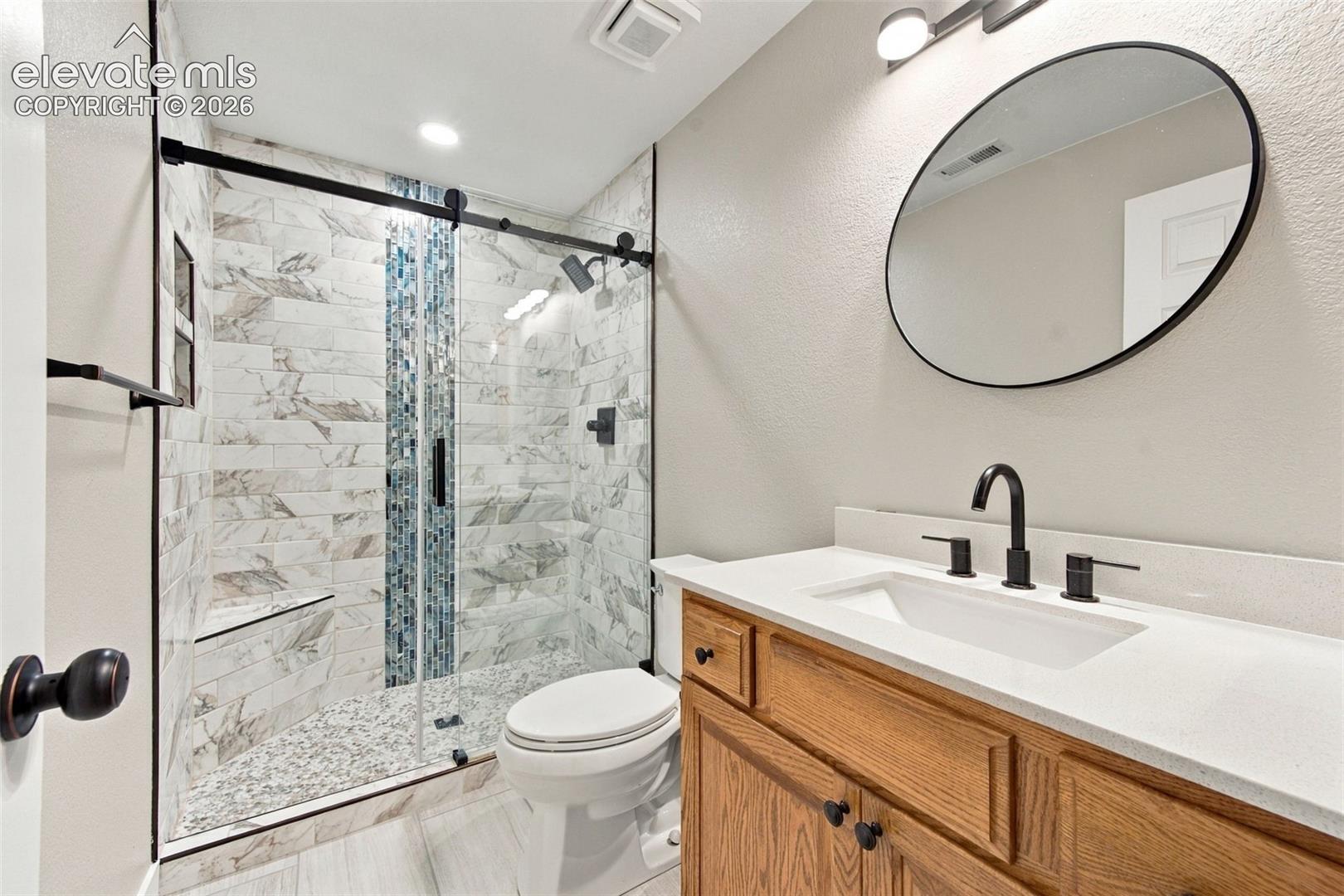 Bathroom featuring a vanity with a light-tone countertop, matte black faucet, and oval wall mirror, complemented by a walk-in shower with a glass sliding door, decorative mosaic tile inlay, and built-in bench seating