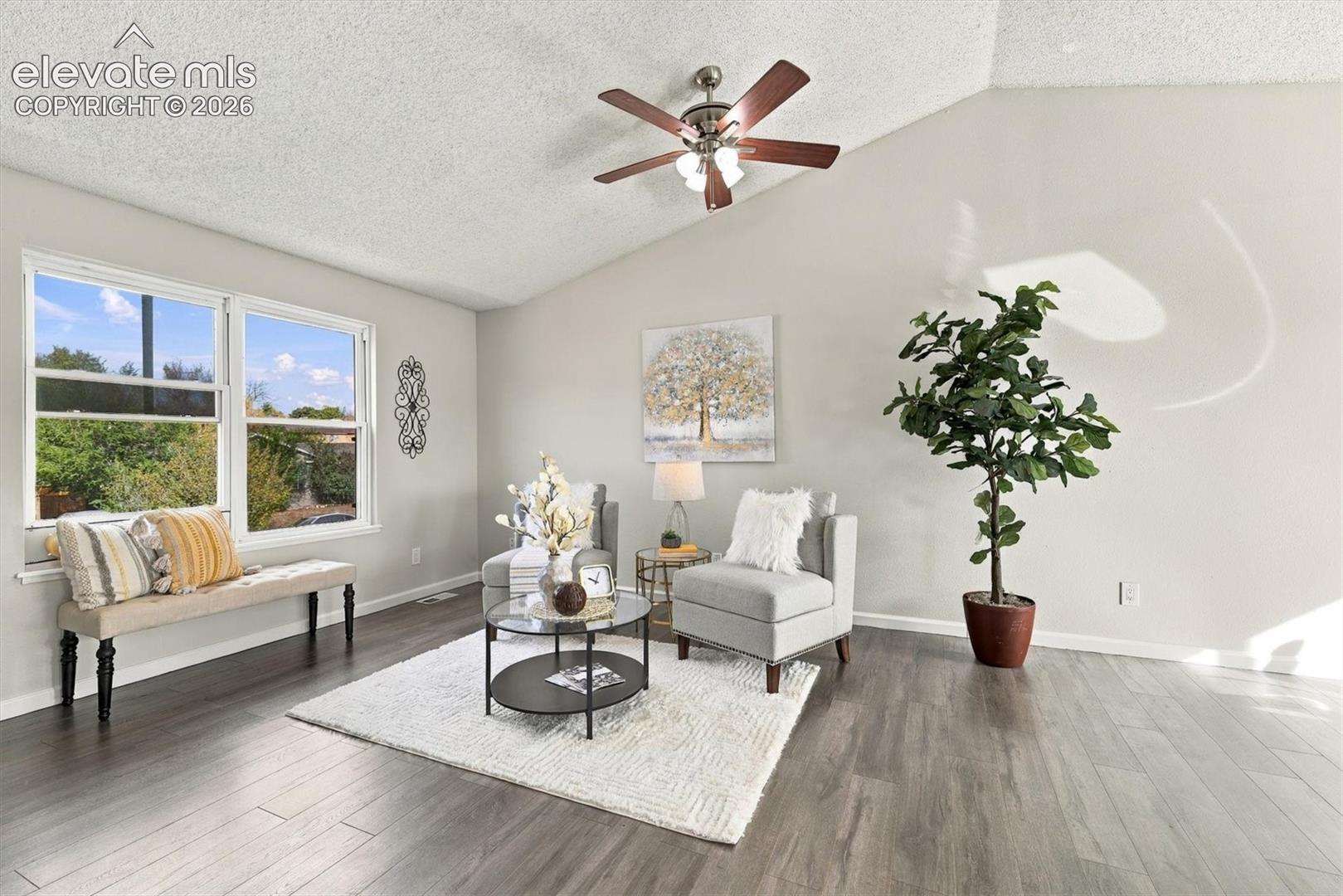Vaulted ceiling featuring a central five-blade ceiling fan with integrated lighting, wood-finish flooring, and large windows providing natural light