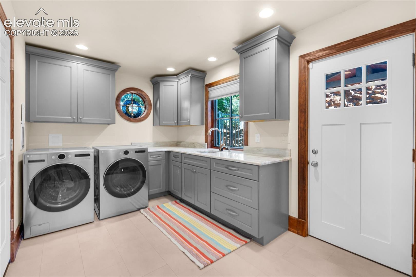 Laundry room featuring grey shaker-style cabinetry, stone countertops, a stainless steel undermount sink, a circular accent window, and a white exterior door with window panes