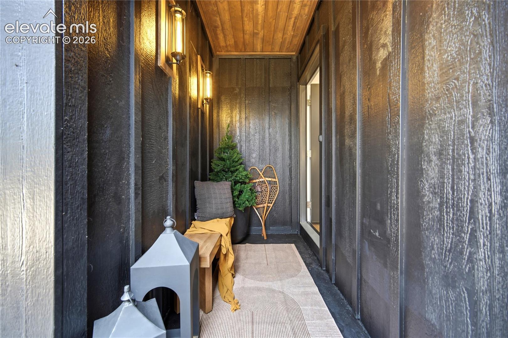 Covered entryway featuring vertical dark wood-finish siding, recessed wood-plank ceiling, and dual wall-mounted cylindrical light fixtures