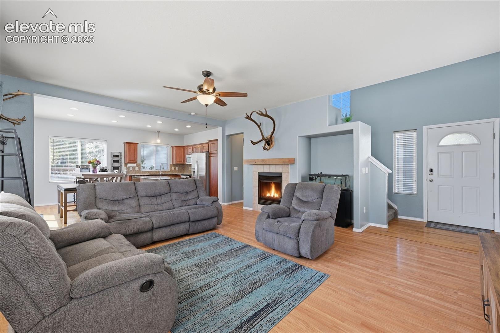 Hardwood flooring throughout, a gas fireplace with a wood mantel, a ceiling fan, and an open-concept design to the kitchen