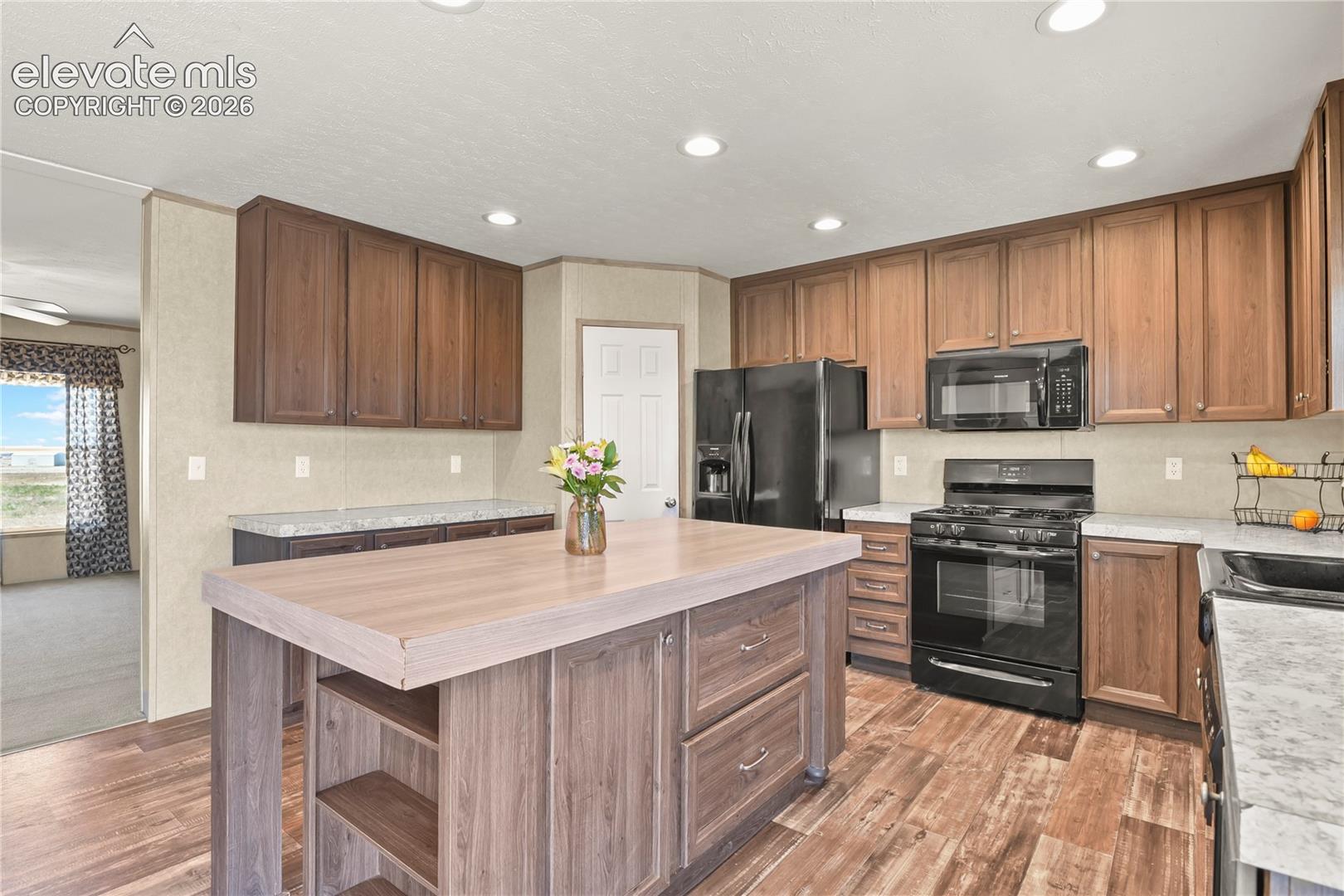 Kitchen featuring stained wood cabinetry, a central island with integrated storage, stone-patterned countertops, wood-finish flooring, and black appliances