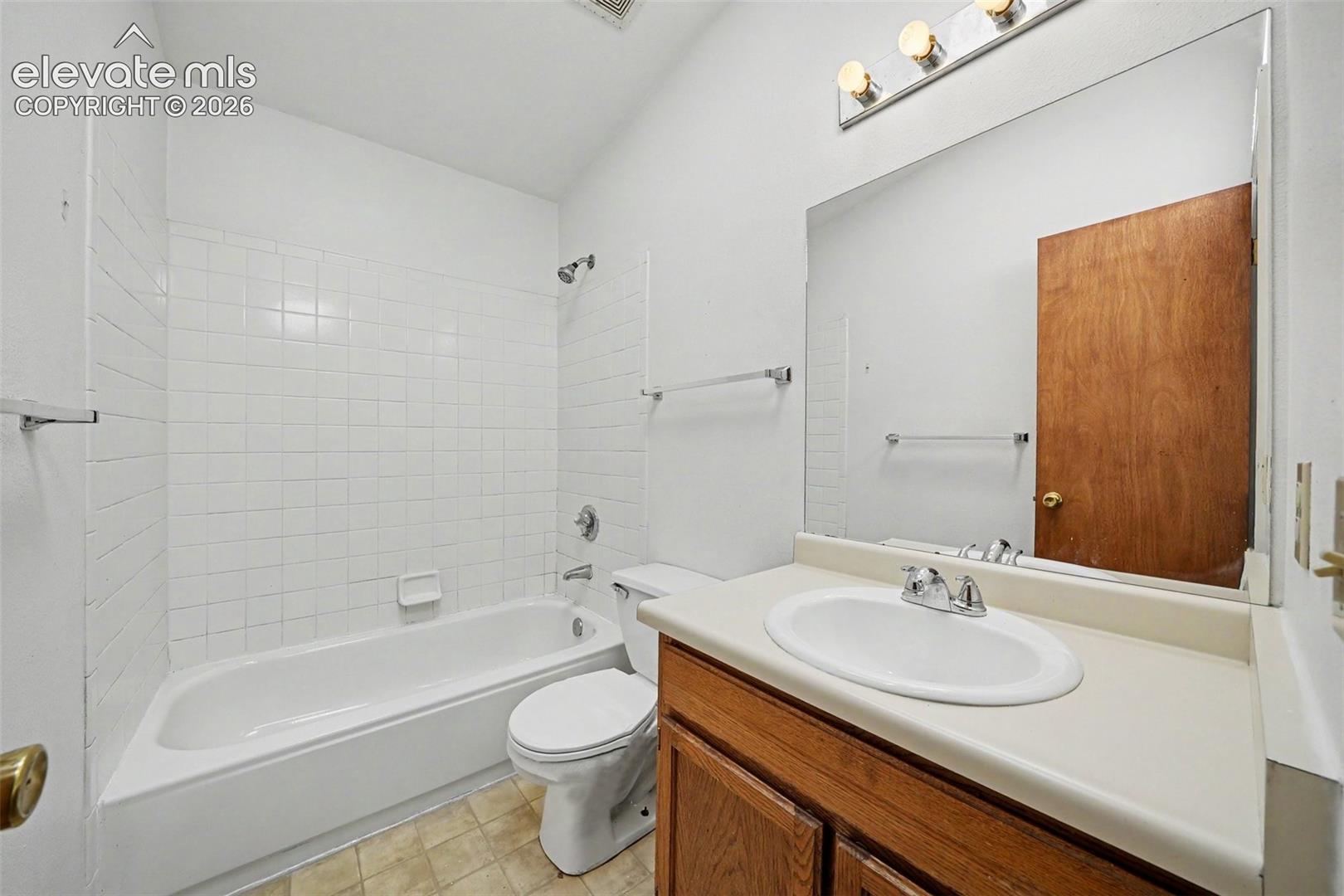 Bathroom featuring a tub-shower combination with white ceramic wall tile, a vanity with a solid-surface countertop and integrated sink, a large wall-mounted mirror, and three-bulb vanity lighting