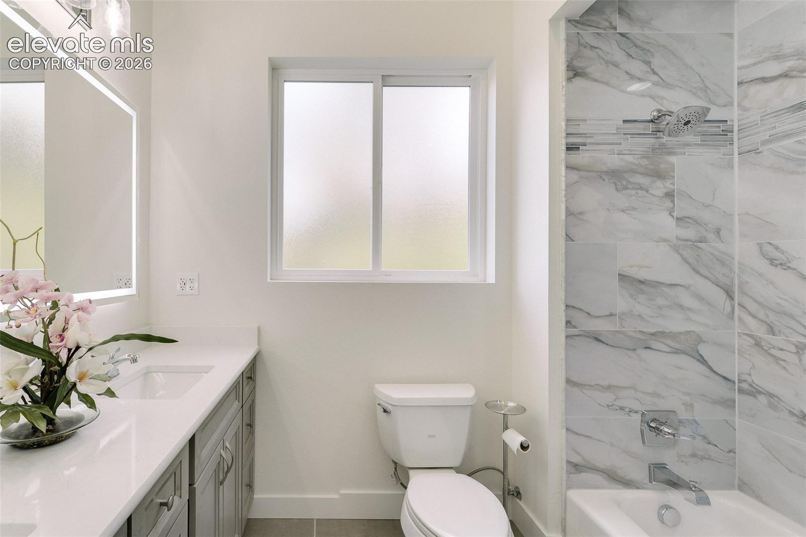 Main Level Full Bathroom with dual sink vanity, quartz countertop, illuminated and fog resistant mirror, and tiled tub/shower.