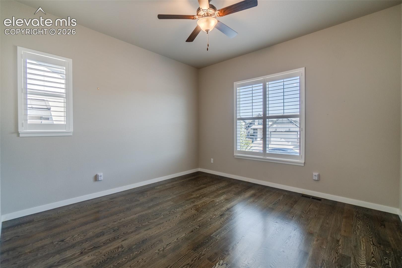 Spacious room featuring wood-finish flooring, two windows with white louvered shutters, a ceiling fan with integrated lighting, and neutral wall paint
