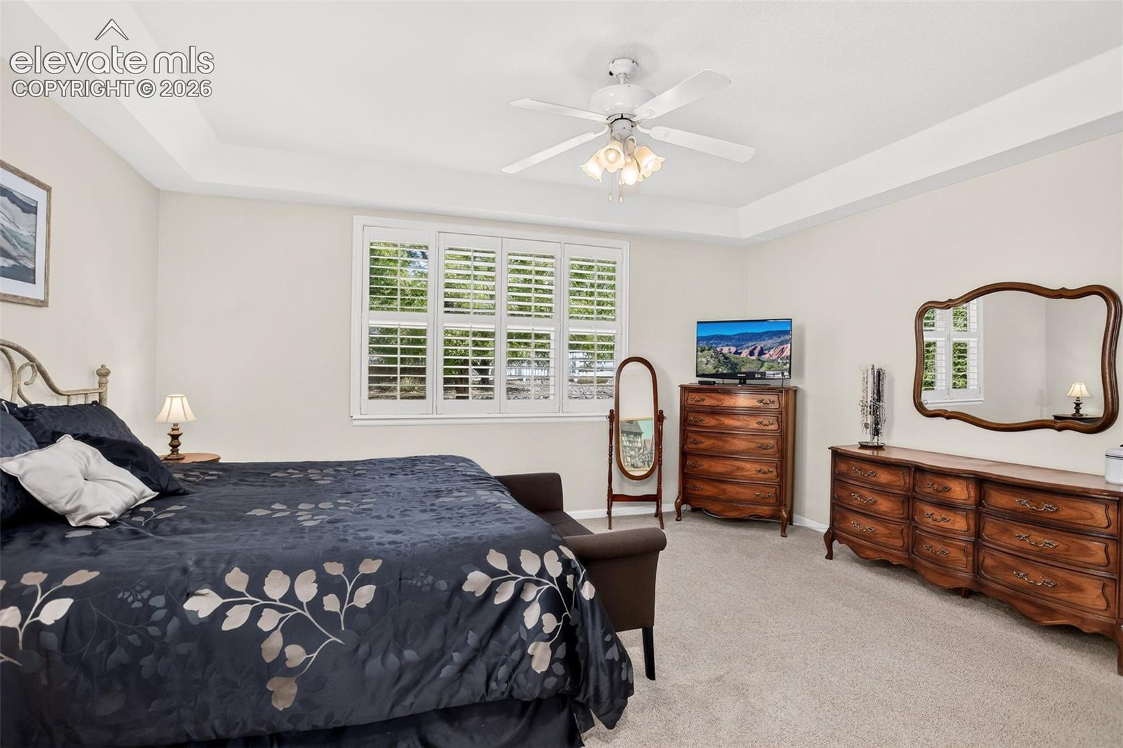 Tray ceiling with recessed lighting, ceiling fan with integrated light fixture, window with white plantation shutters, and light-colored carpeting