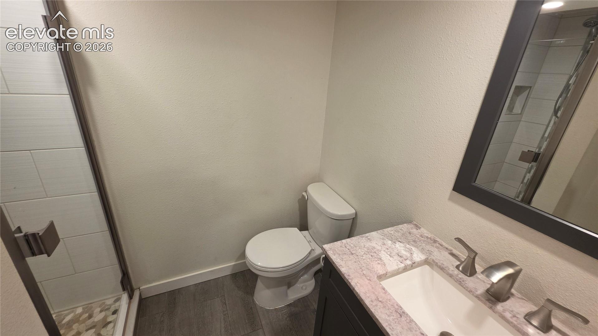 Vanity with integrated sink and brushed nickel faucet, framed mirror, wood-finish flooring, and a glass-enclosed shower featuring tiled walls and a mosaic-patterned base