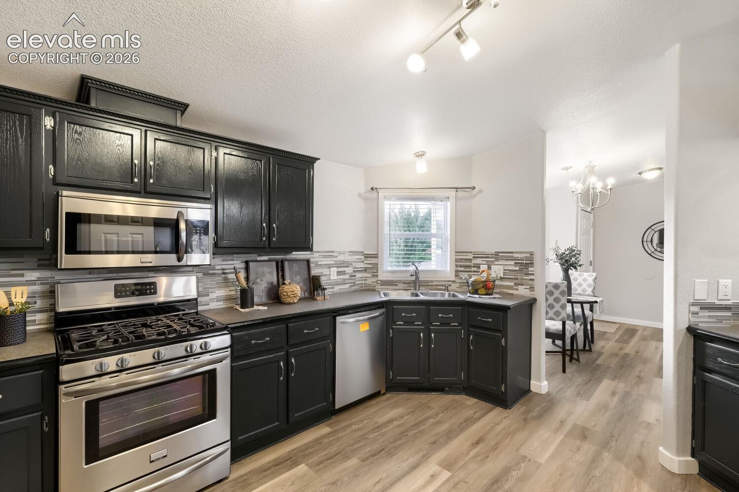 Kitchen featuring dark cabinetry, stainless steel appliances, stone-tile backsplash, wood-finish flooring, and track lighting