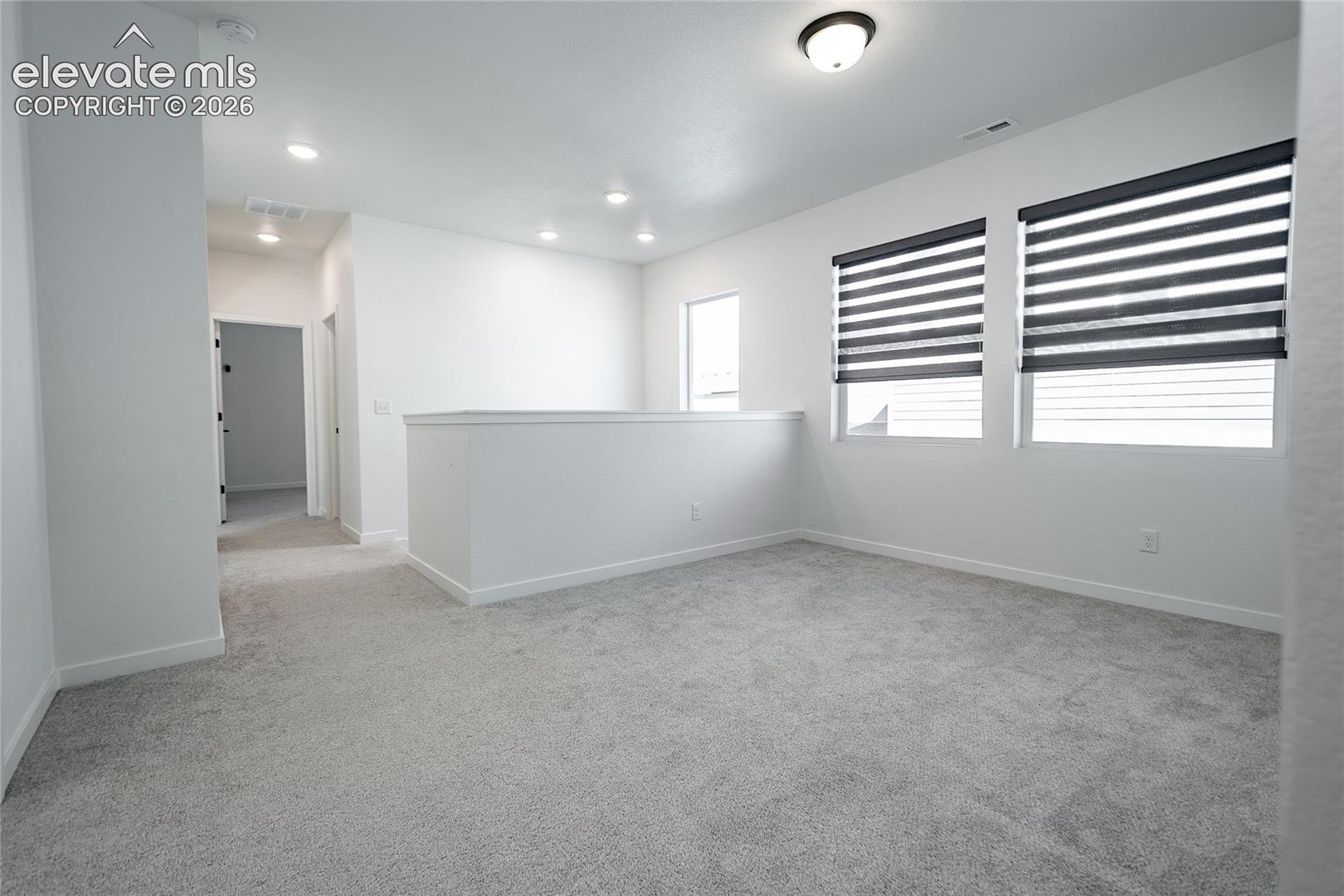 Open space featuring neutral-toned carpeting, recessed ceiling lighting, a flush-mount fixture, and dual windows with installed roller shades