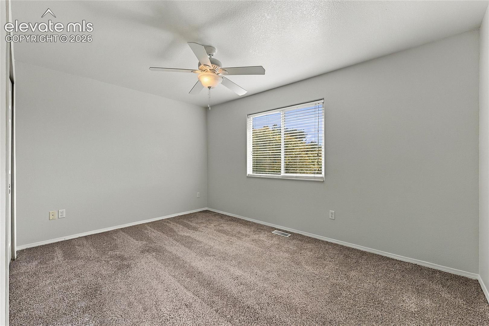 Neutral wall tones, carpeted flooring, sliding window with horizontal blinds, and a ceiling-mounted fan with light fixture