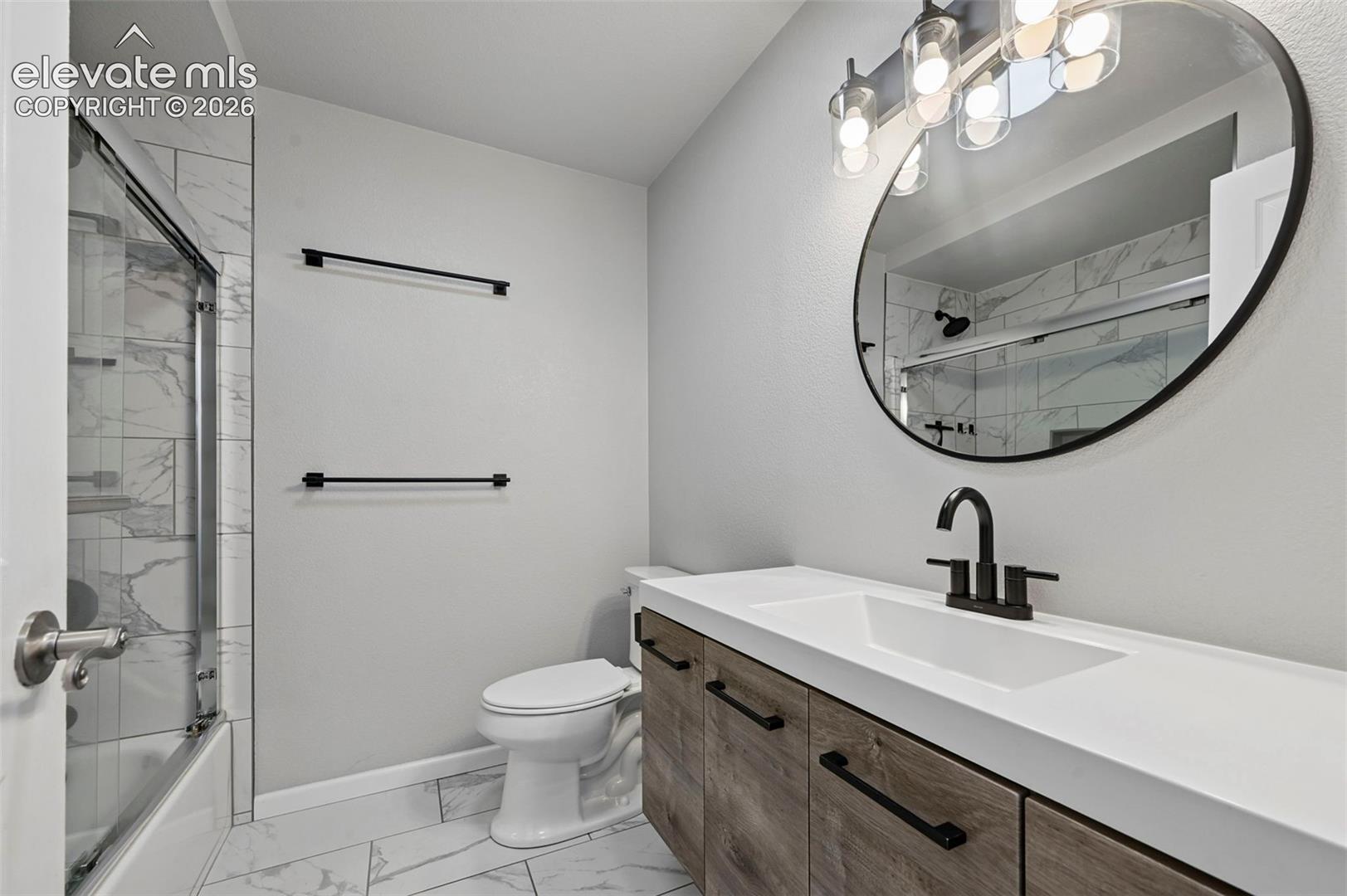 Beautifully remodeled bathroom on main floor, adjacent to secondary bedroom-  with wood-finish cabinetry, white countertop, integrated sink, and matte black hardware, accompanied by an oval mirror with modern lighting, tiled flooring, and a shower-tub combination with light-toned wall tile