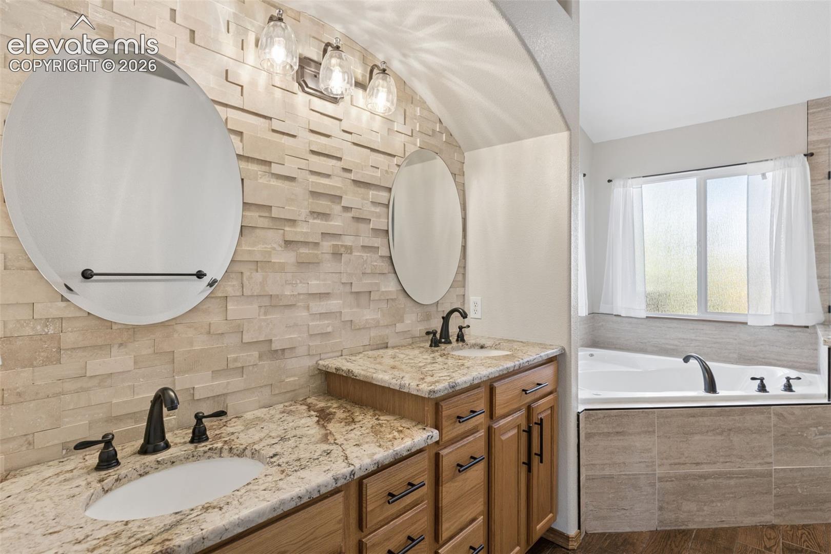 Dual vanity with stone-finish countertops, textured stone backsplash, two circular wall-mounted mirrors, dark finish hardware, and built-in soaking tub
