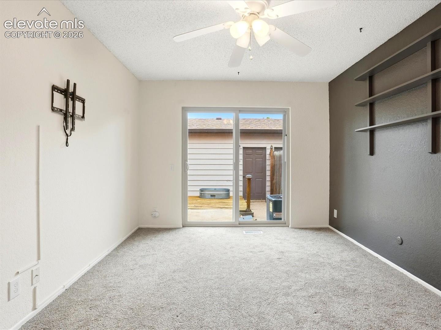 Neutral-toned carpeted flooring, sliding glass door, ceiling-mounted fan with light fixture, and dark-toned accent wall featuring built-in shelving