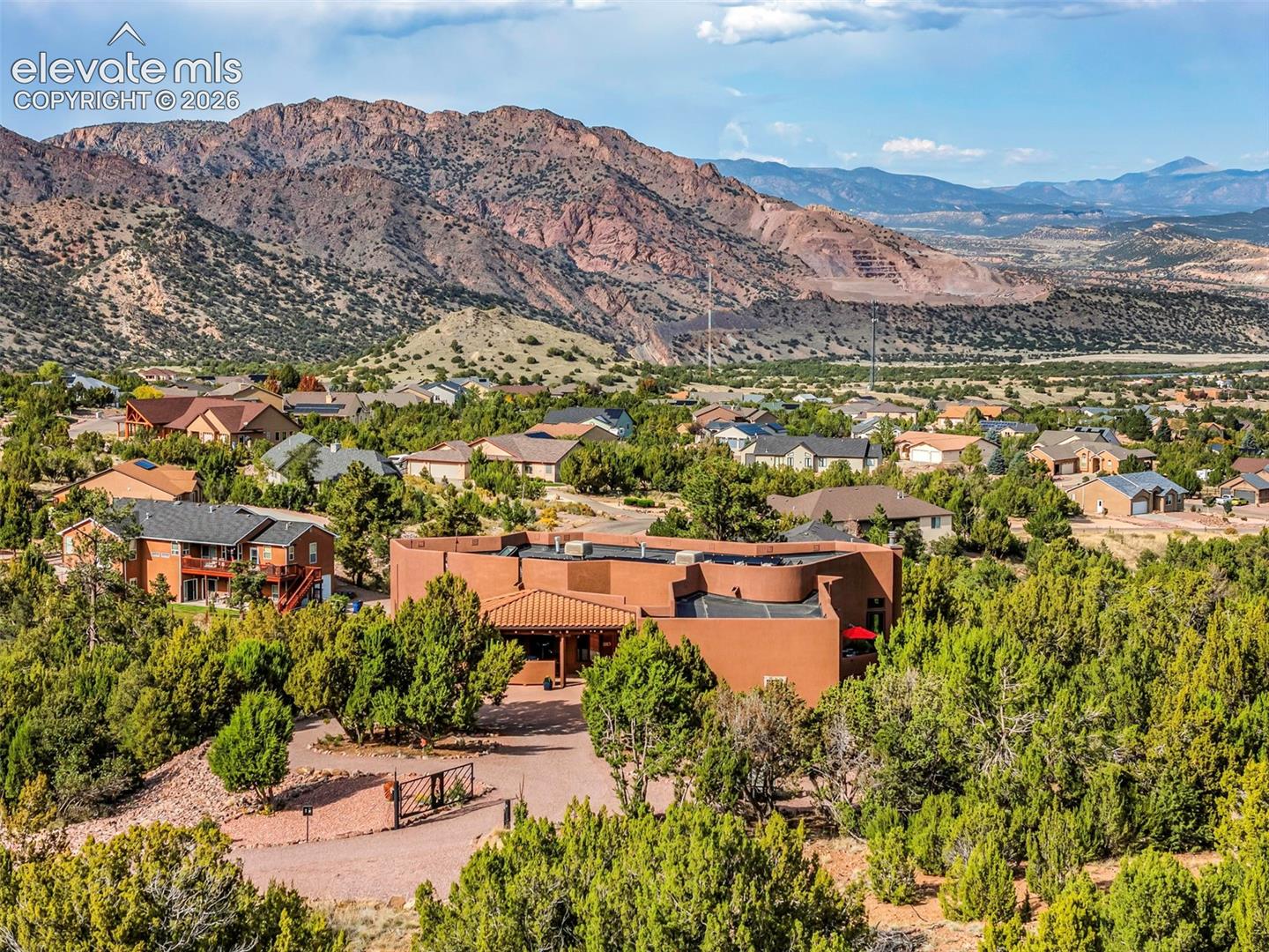 Stucco exterior residence featuring a flat roof with tile accents, a gravel driveway, and a gated entrance, set against a mountainous landscape