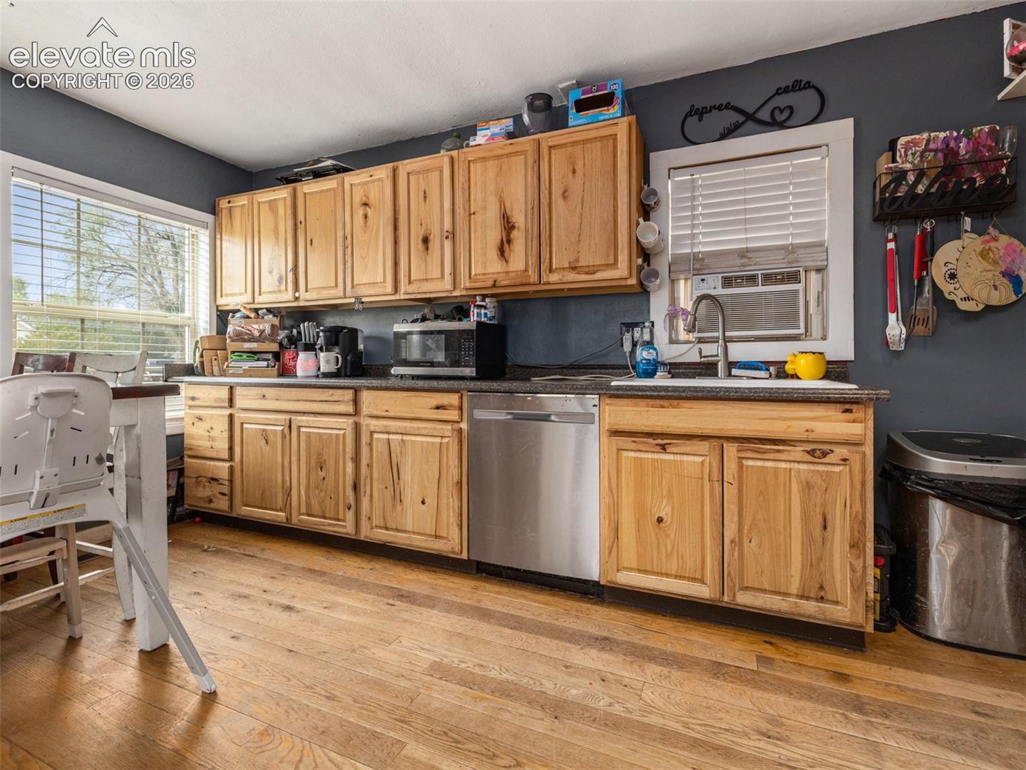 Spacious kitchen featuring warm wood-finish cabinetry with ample storage, stainless steel appliances including a dishwasher, dark countertops with workspace, wood-finish flooring throughout, and natural light from multiple windows