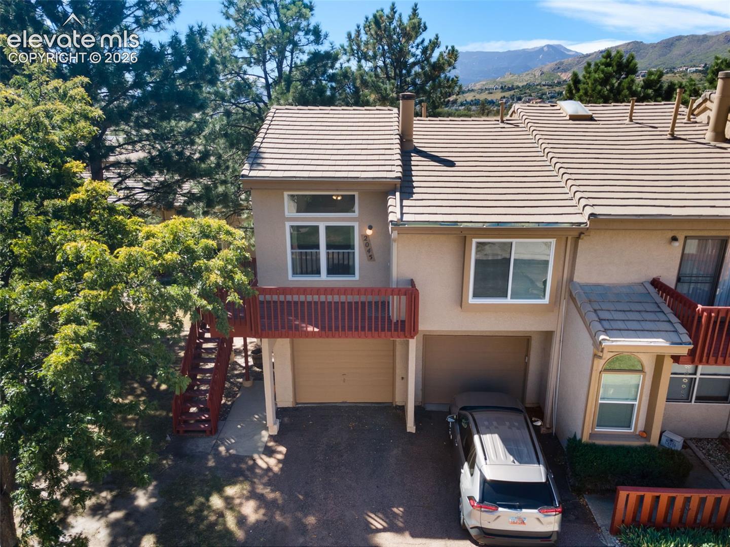 Exterior facade featuring stucco siding, a tiled roof, a two-car garage, an elevated wooden balcony with integrated stairs, and mountain backdrop