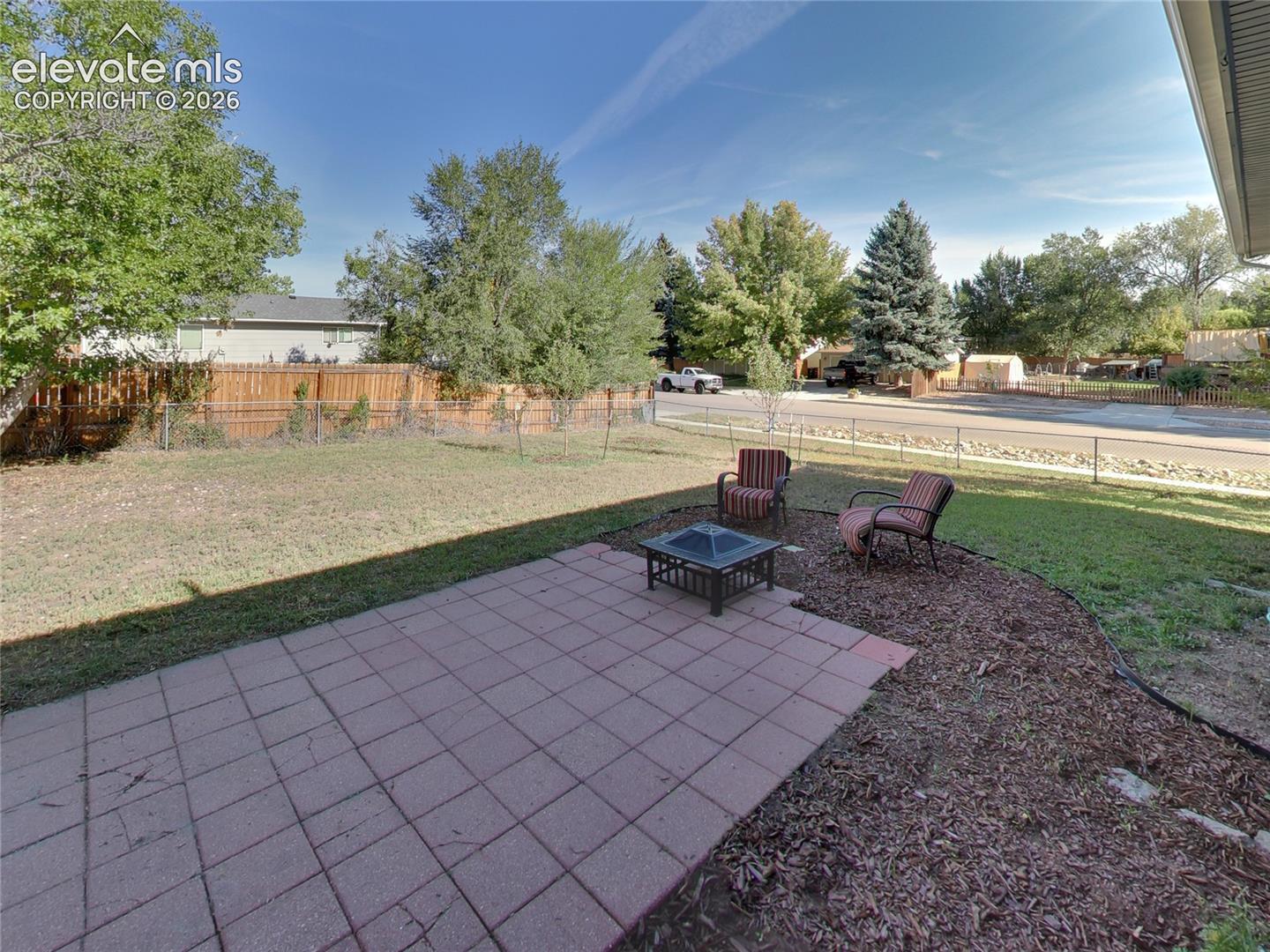 Paved patio area, expansive lawn, perimeter fencing, mature trees, and a landscaped border