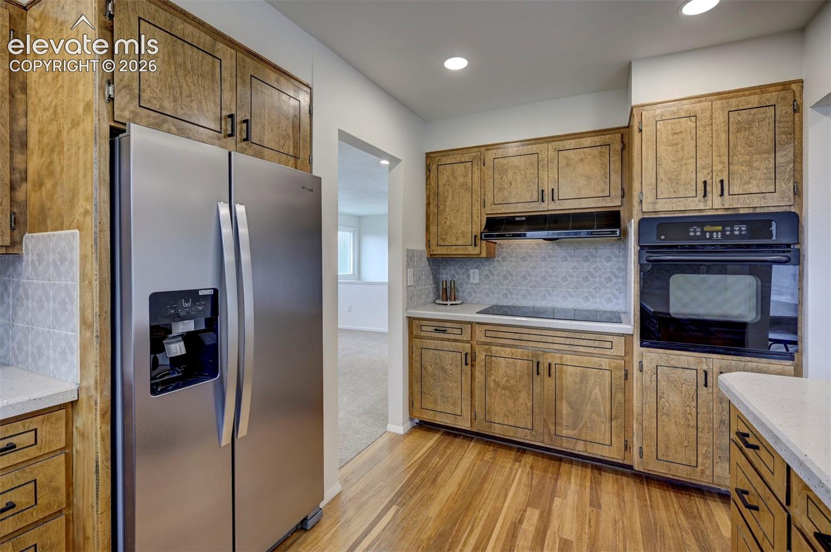 Kitchen featuring wood-finish cabinetry, stainless steel side-by-side refrigerator, built-in oven, flat-surface cooktop, and light-toned solid surface countertops