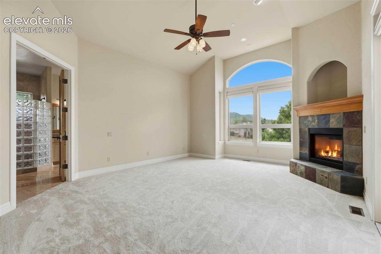 Spacious room featuring vaulted ceilings, an arched window with expansive outdoor scenery, a corner fireplace with a wood mantle and stone surround, and light-toned carpeting