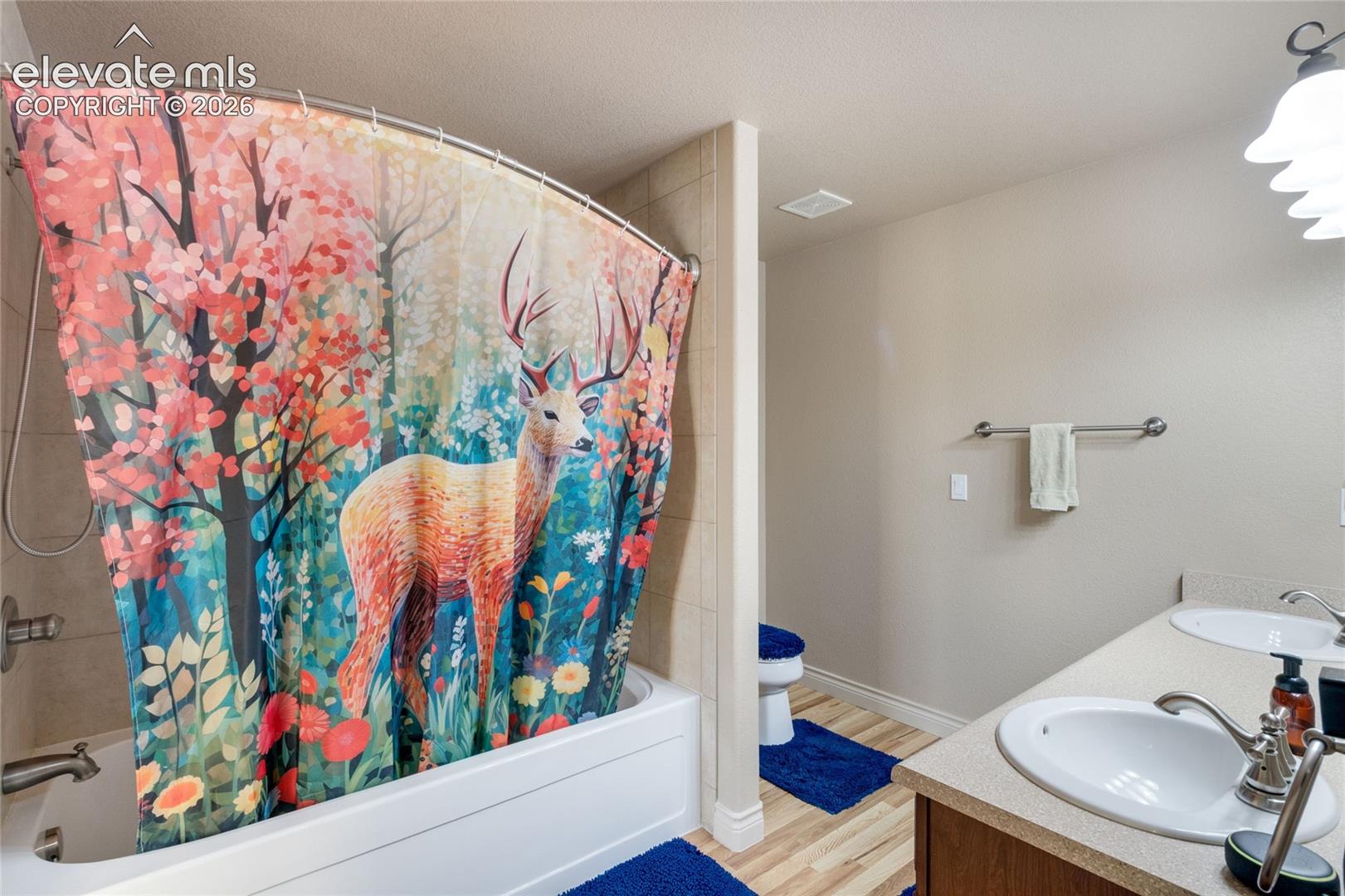 Basement Bathroom featuring a dual-sink vanity with light-toned solid surface countertops, polished chrome fixtures, tile surround bathtub, and wall-mounted multi-bulb lighting