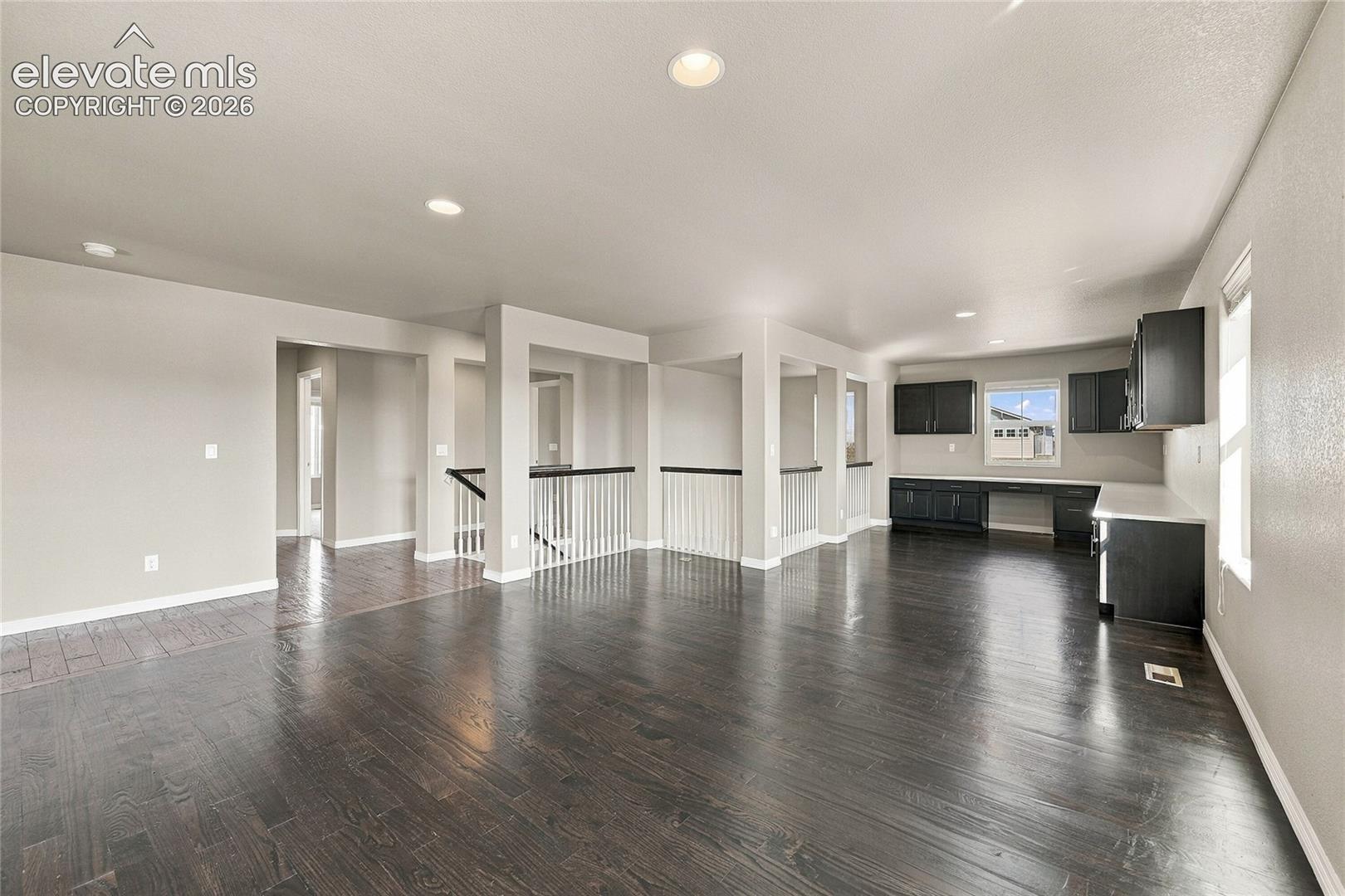 Open-concept interior featuring wood-finish flooring, recessed lighting, built-in dark cabinetry with solid-surface countertops, and a white balustrade railing