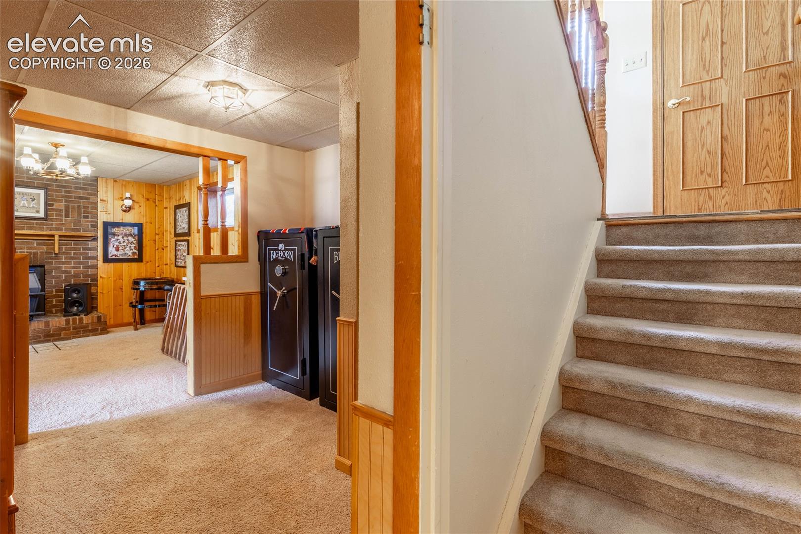 Interior featuring wood-paneled walls, a brick fireplace, drop ceiling, and carpeted staircase with wood handrail