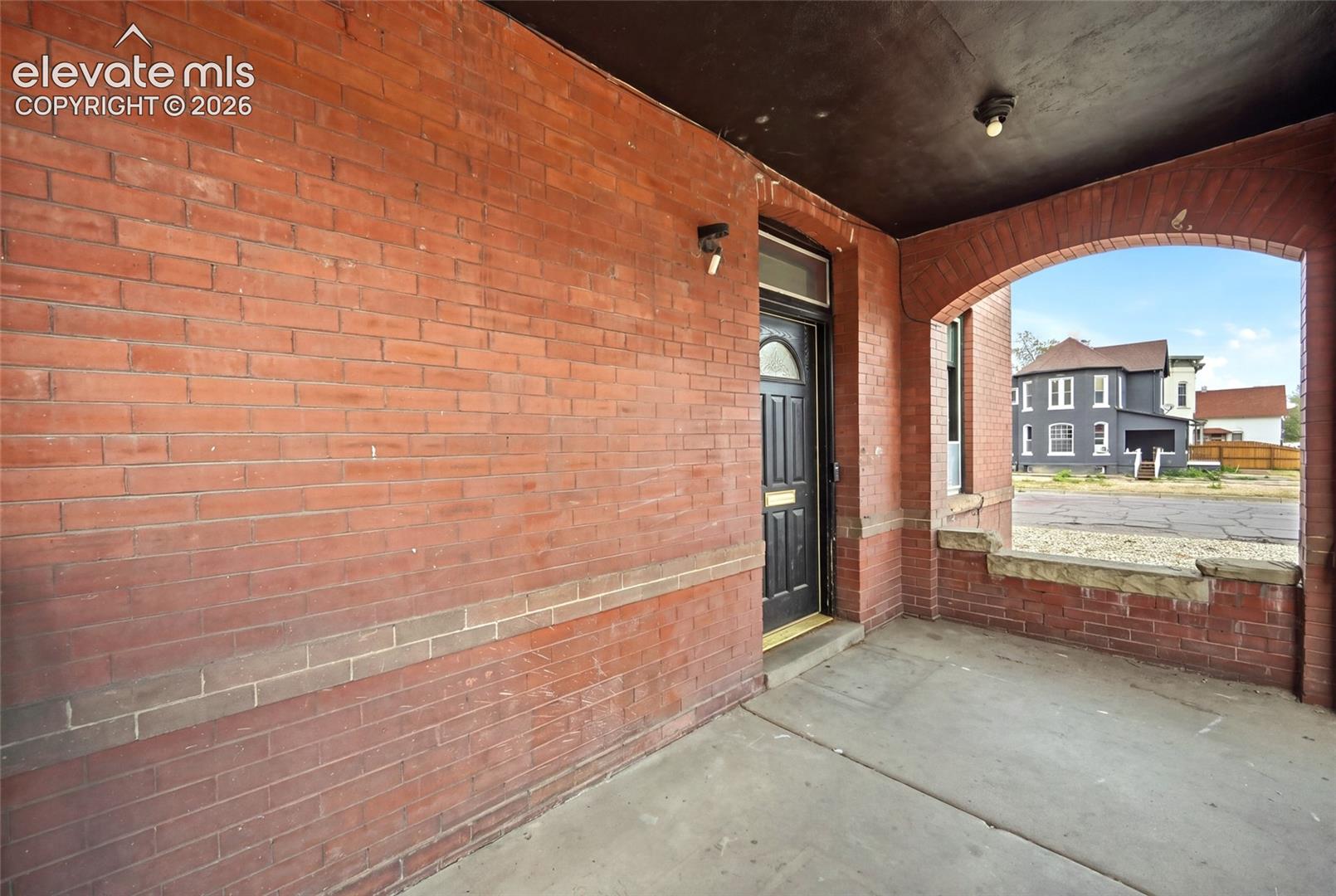 Covered front porch featuring exposed red brick masonry, a concrete floor, an arched entry, and a paneled entry door with a mail slot