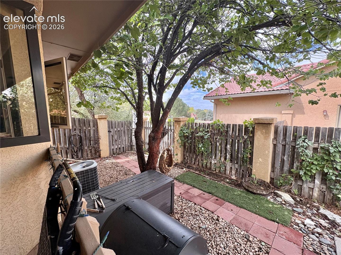 Covered patio space featuring a mature tree, privacy fencing, and a gravel and brick paver pathway