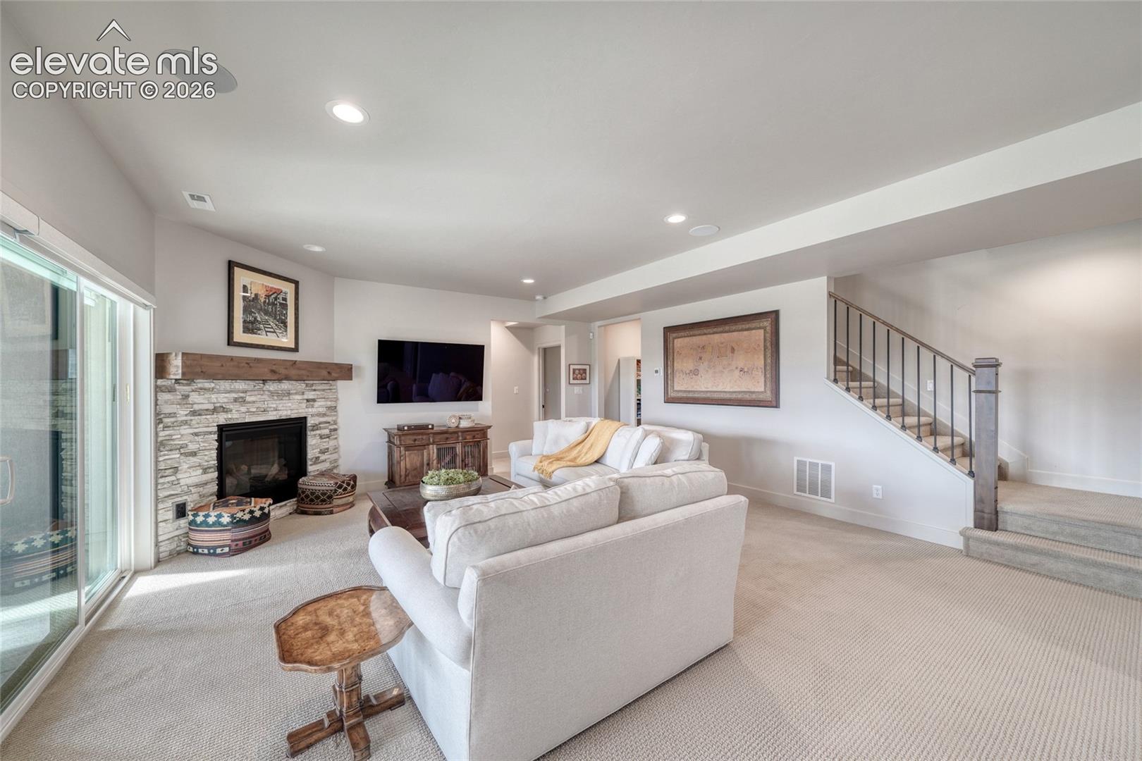 Open-concept living space featuring a floor-to-ceiling stone-veneer fireplace with a heavy timber mantel, recessed ceiling lighting, carpeted flooring, and a staircase with decorative metal balusters