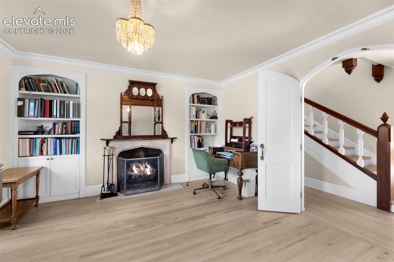 Fireplace with stone surround and integrated mirror, dual arched built-in bookcases, wood-finish flooring, crystal chandelier, and arched doorway with wooden staircase