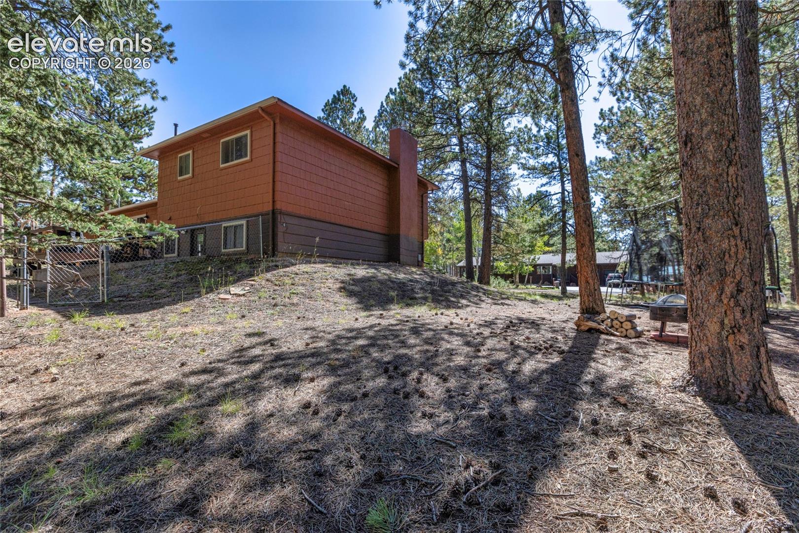 Wood-sided exterior with a natural, sloped landscape featuring mature trees and a chain-link fence