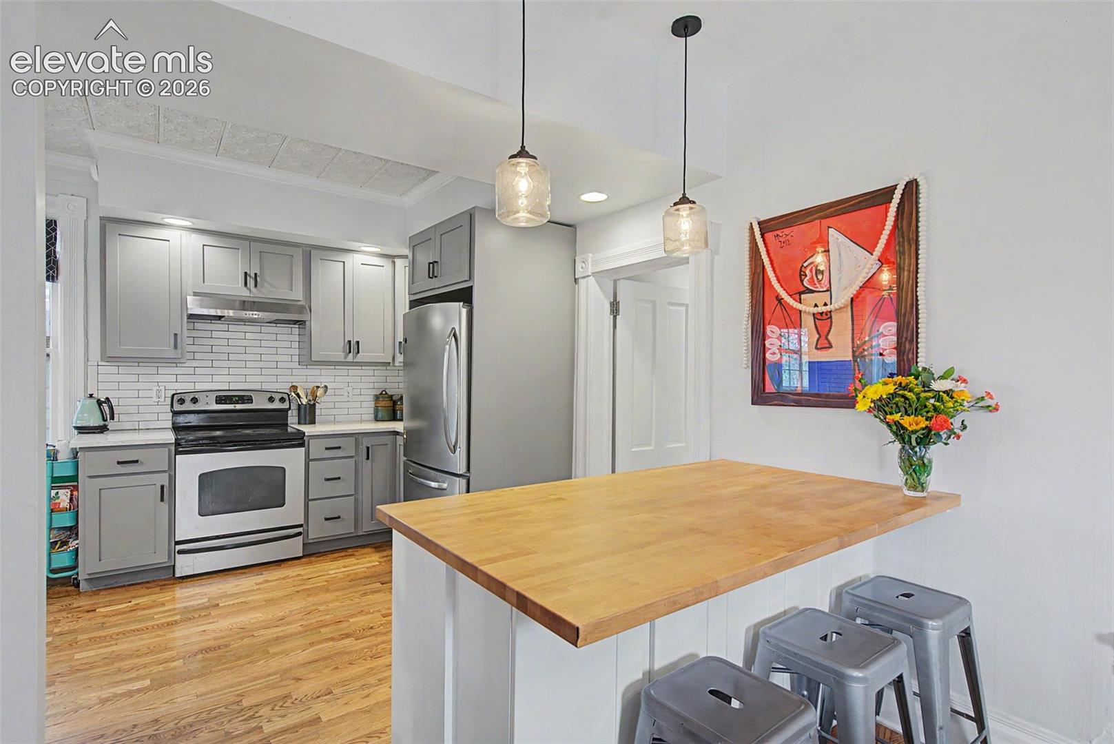 Updated kitchen featuring light gray cabinetry, white tile backsplash, stainless steel appliances, a wood-finish breakfast bar/ kitchen island, and hardwood flooring