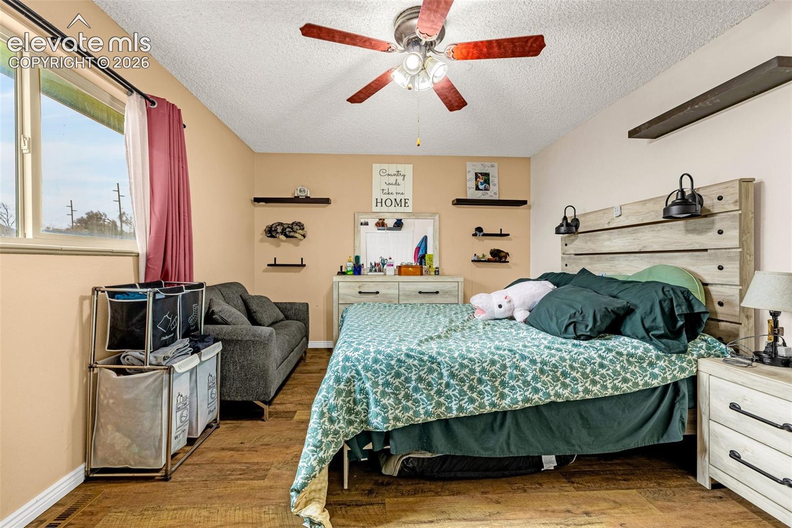 Bedroom featuring wood-finish flooring, a ceiling fan with integrated lighting, wall-mounted floating shelving, and a horizontal wood-plank headboard with mounted sconces